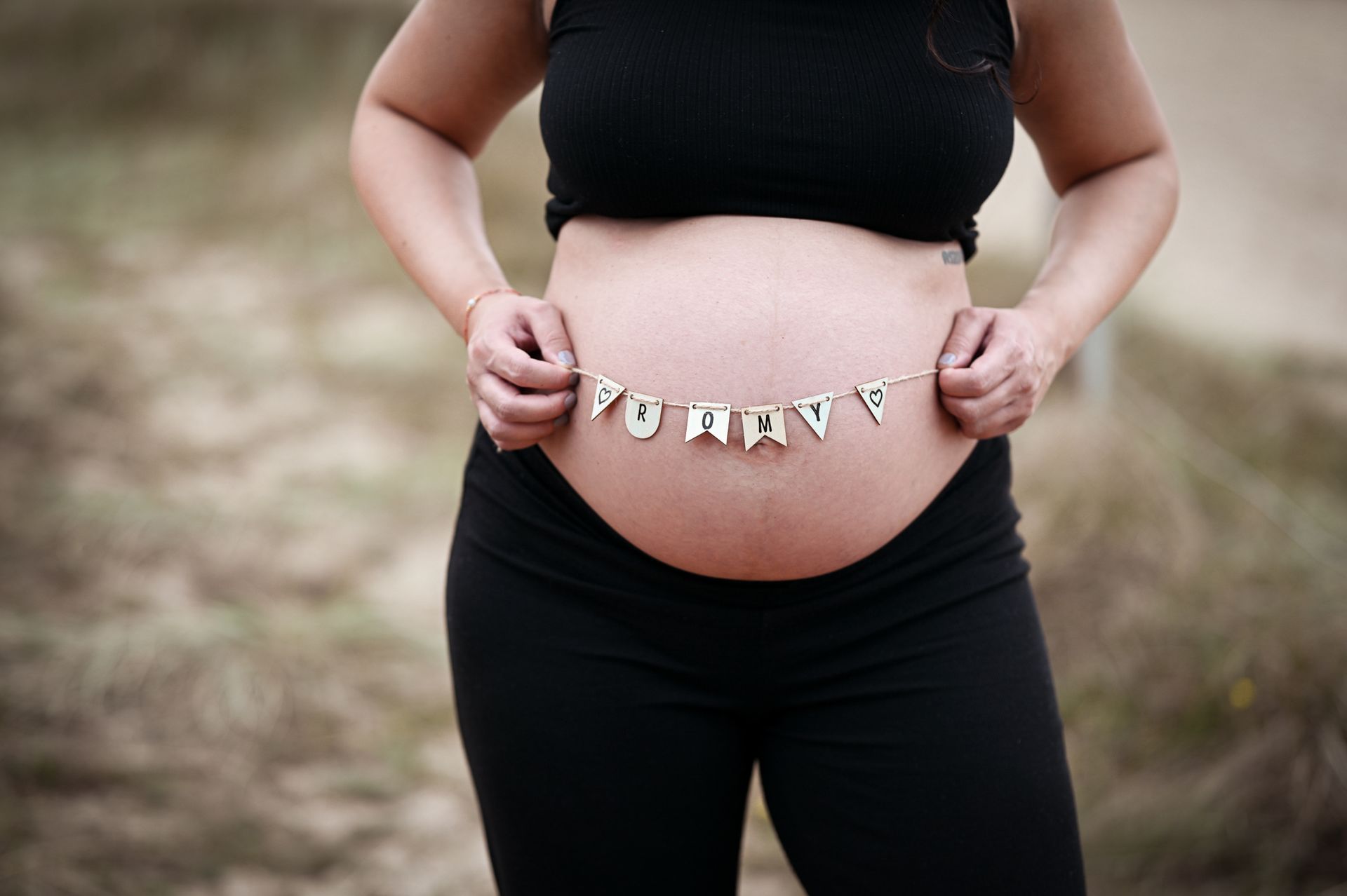 Schwangere Person hält ein Banner mit der Aufschrift „Mami“ über ihrem Bauch. Sie trägt schwarze Kleidung und steht im Freien. Kirstin Hammerstein, hammerstein-pictures