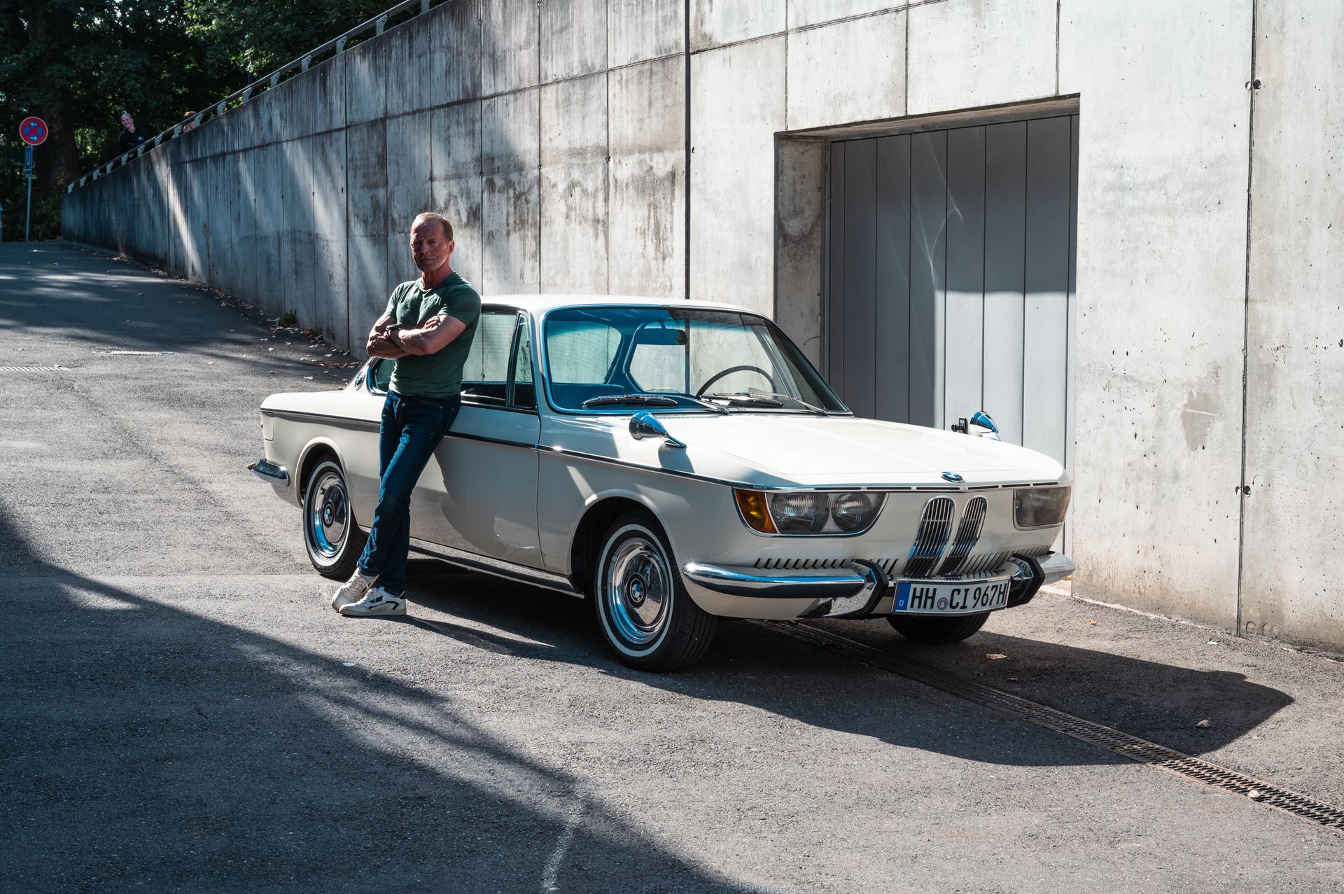 Mann in Jeans lehnt auf einer Stadtstraße an einem klassischen weißen BMW. Betonwand im Hintergrund. Kirstin Hammerstein, hammerstein-pictures