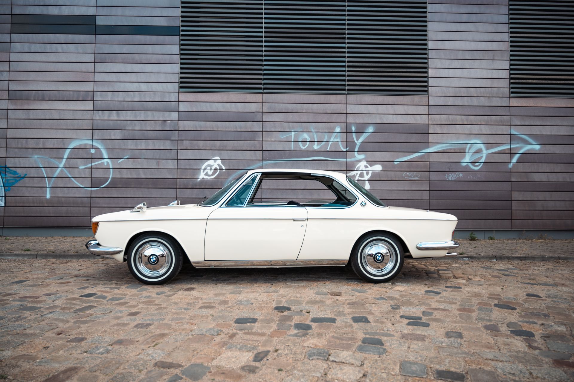 Weißer Oldtimer, der auf einer Kopfsteinpflasterstraße mit einer mit Graffiti bedeckten Wand im Hintergrund geparkt ist. Kirstin Hammerstein, hammerstein-pictures