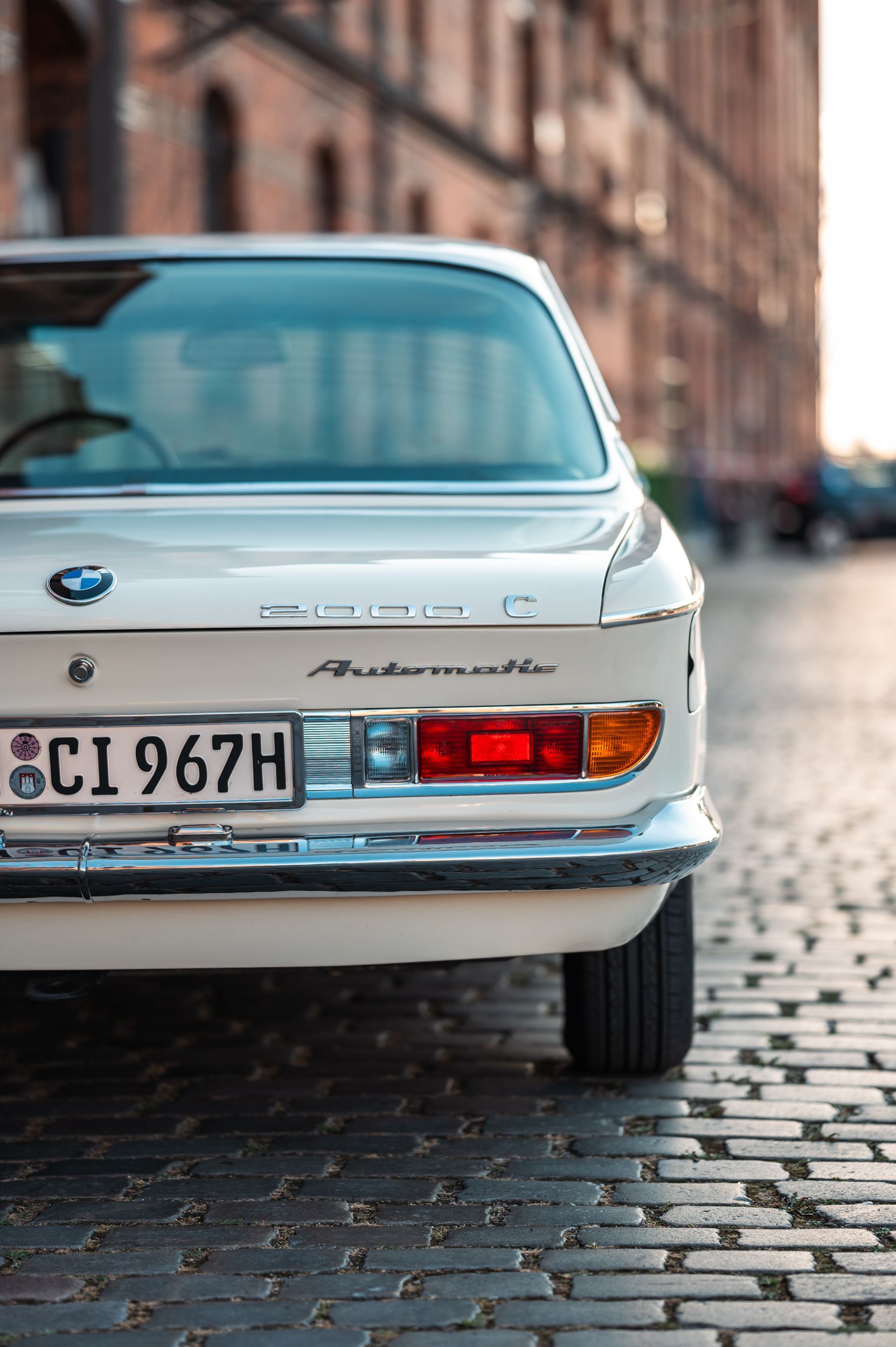 Rückansicht eines weißen Vintage-BMW-Coupés, das auf Kopfsteinpflaster geparkt ist, mit Rücklichtern, Nummernschild und Gebäudehintergrund. Kirstin Hammerstein, hammerstein-pictures
