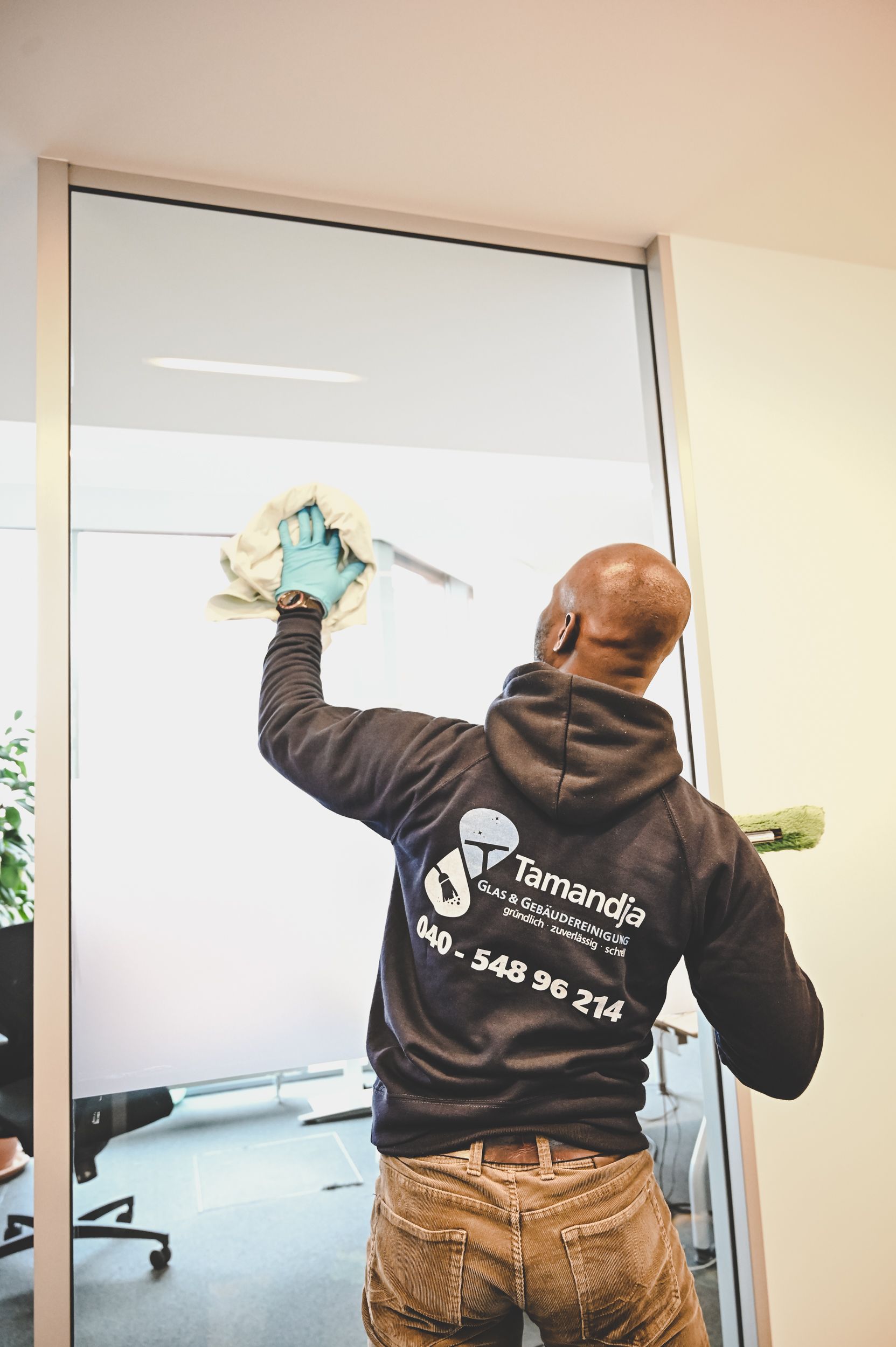 Kirstin Hammerstein, hammerstein-pictures. Ein Mann reinigt eine Glastrennwand im Büro mit einem Tuch und einem Abzieher; er trägt einen Kapuzenpullover und Handschuhe.