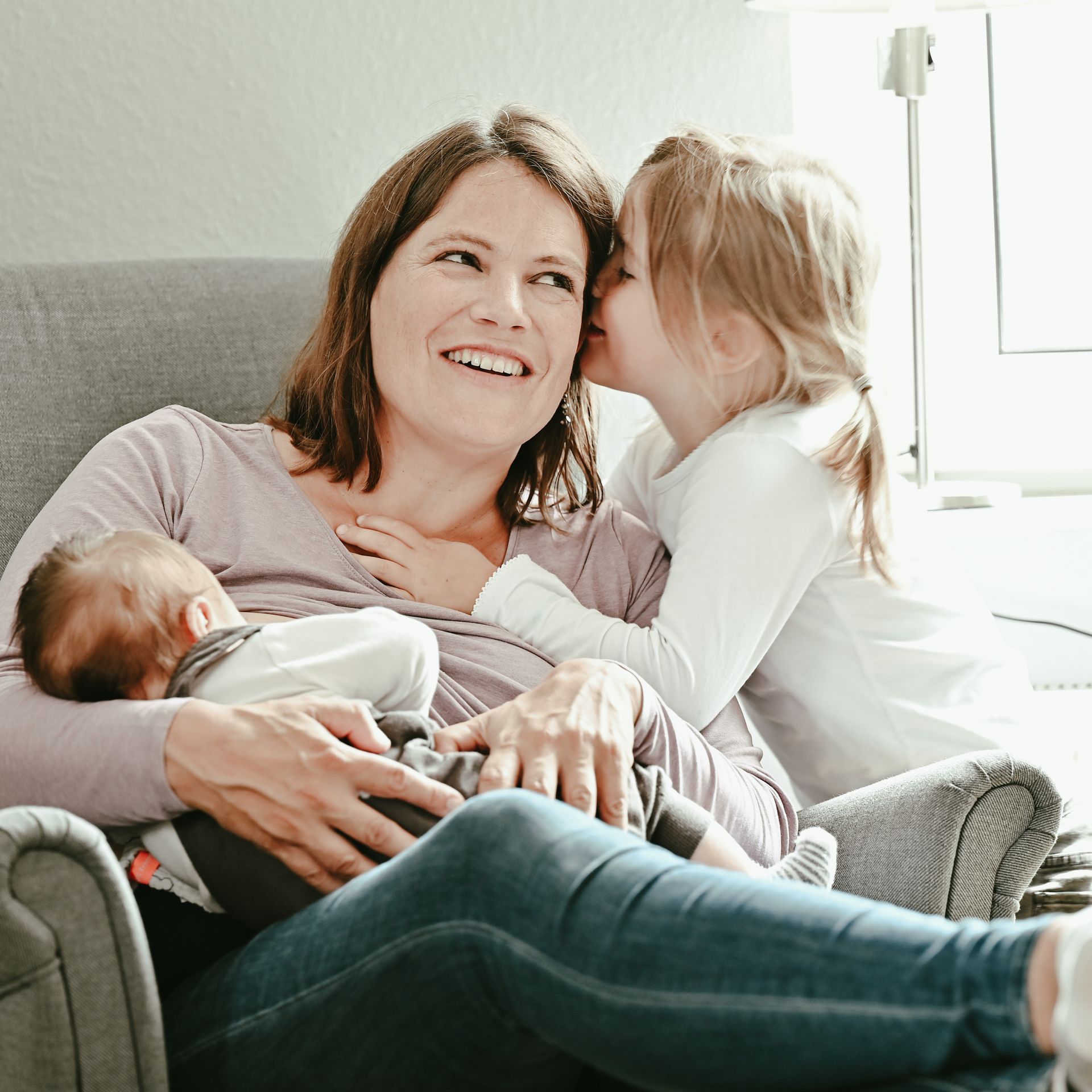 Lächelnde Frau, die ihr Baby stillt, während ihr das kleine Kind etwas ins Ohr flüstert. Innenaufnahme, helle Farben. Kirstin Hammerstein, hammerstein-pictures