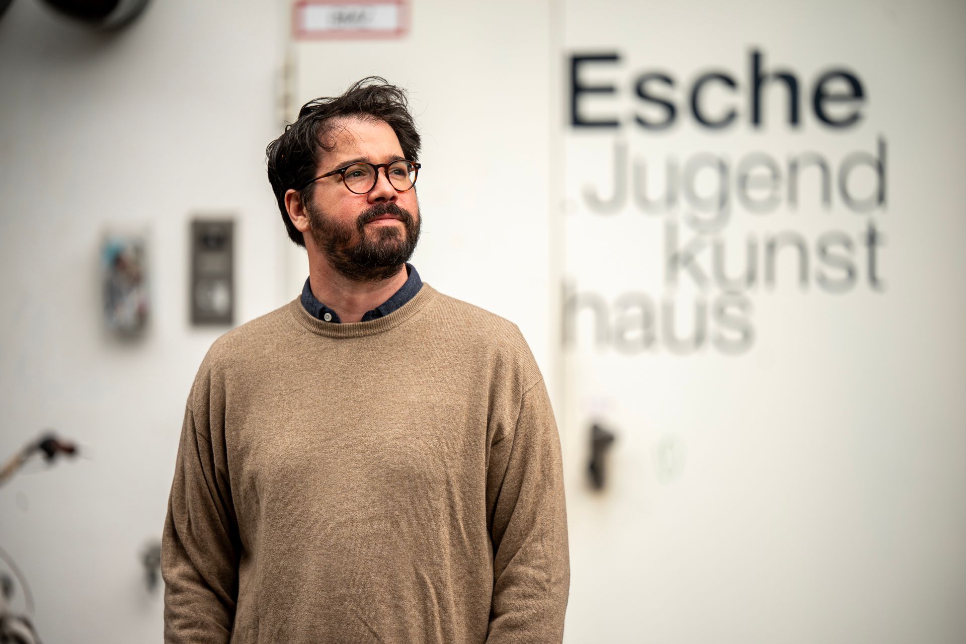 Ein Mann mit Brille und Pullover steht vor einer weißen Wand mit der Aufschrift „Escher Jugend Kunsthaus“.