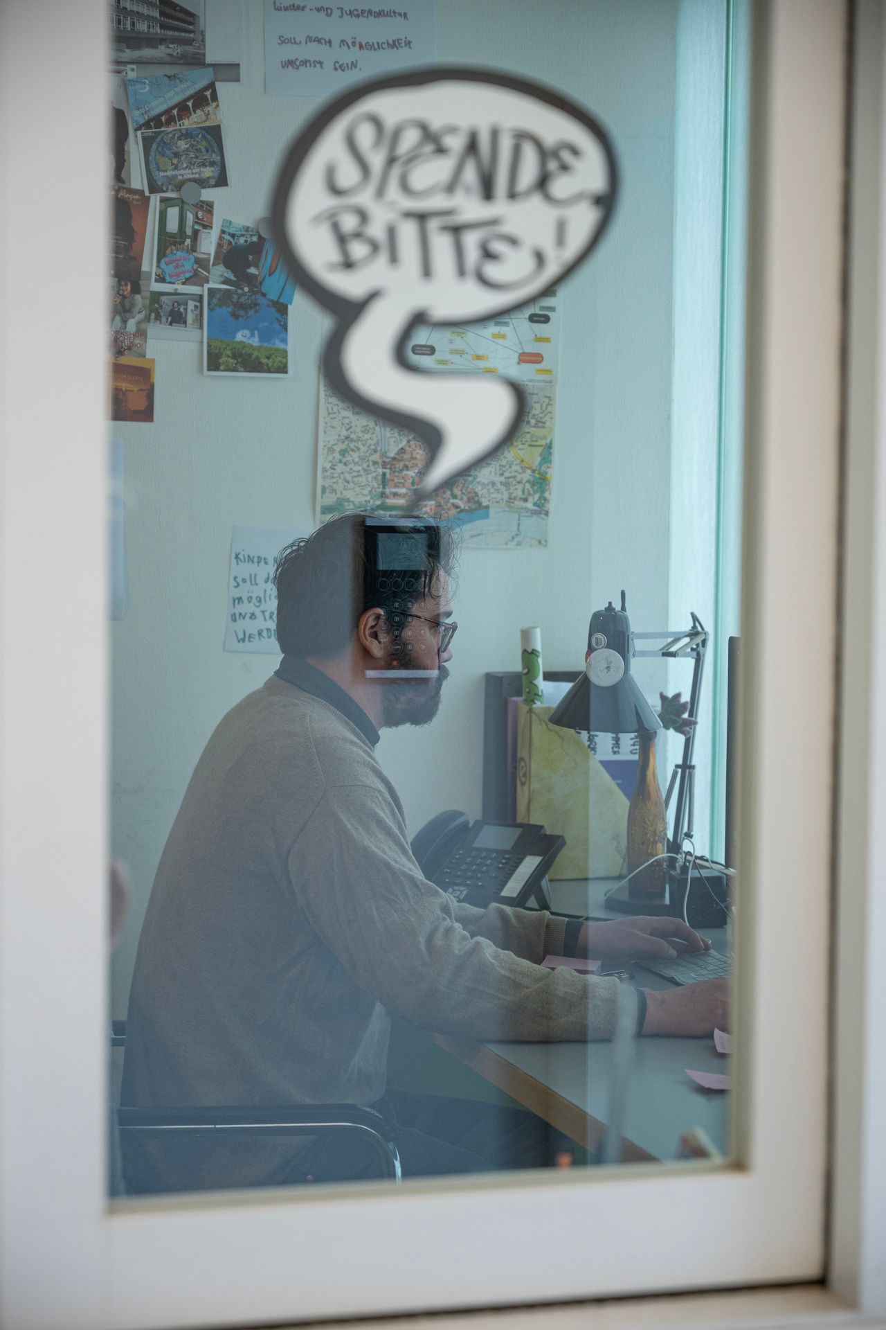 Eine Person sitzt an einem Schreibtisch und arbeitet an einem Computer hinter einer Glasscheibe mit einem Aufkleber mit der Sprechblase „Spende Bitte“.