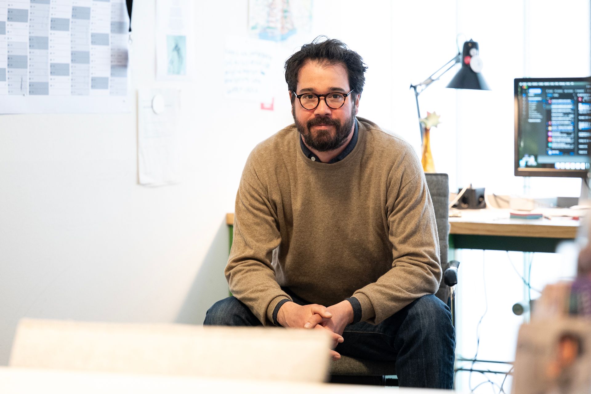 Mann mit Bart und Brille sitzt in einem Pullover und schaut in die Kamera. Büroszene.