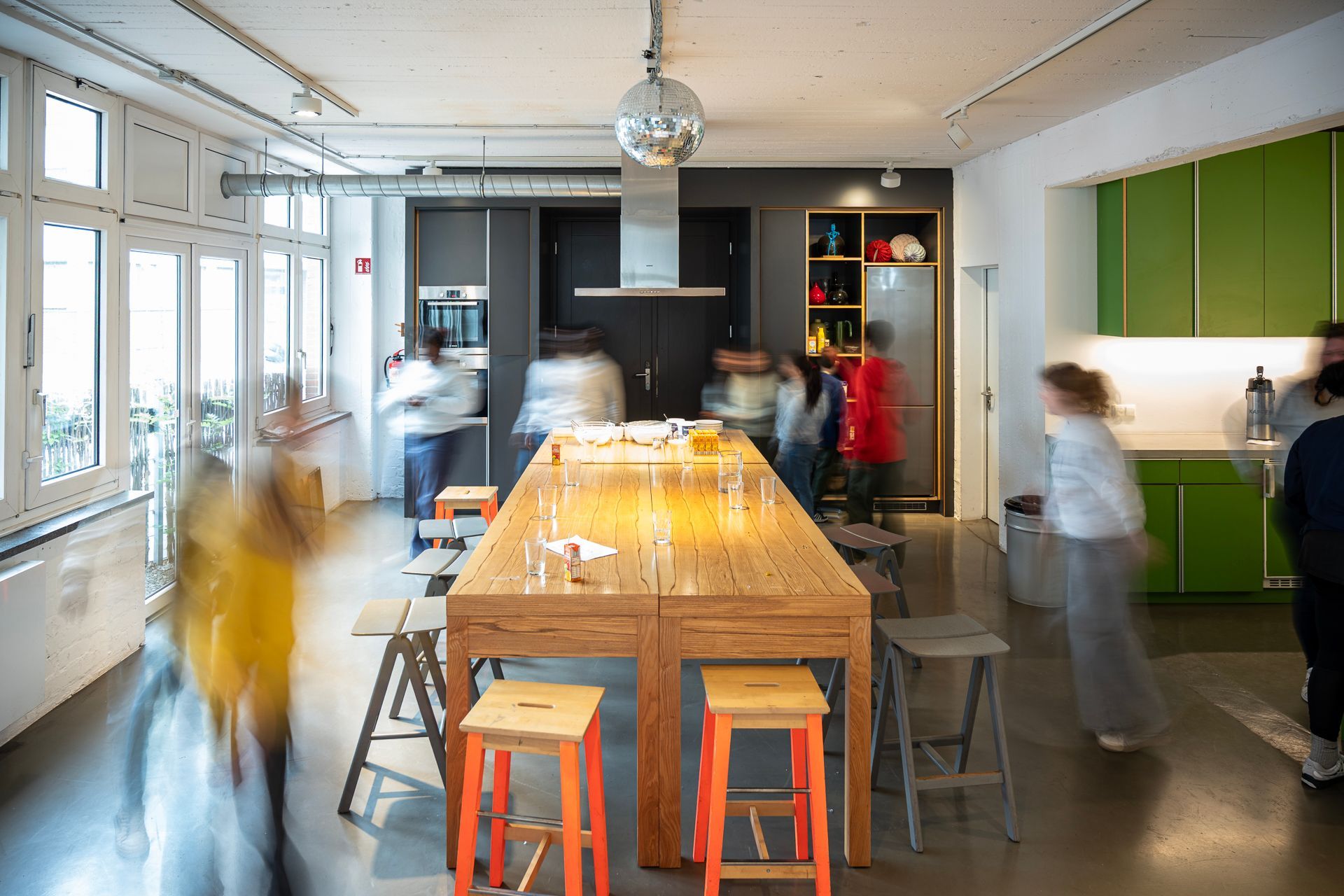 Eine hell erleuchtete Küche mit einem langen Tisch, Hockern und verschwommenen Gestalten von Menschen, die sich dort bewegen.
