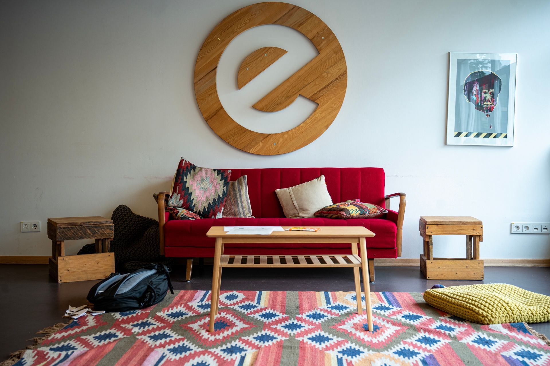 Gemütliches Zimmer mit rotem Sofa, Holztisch und buntem Teppich. An der Wand prangt ein großes „e“-Logo.