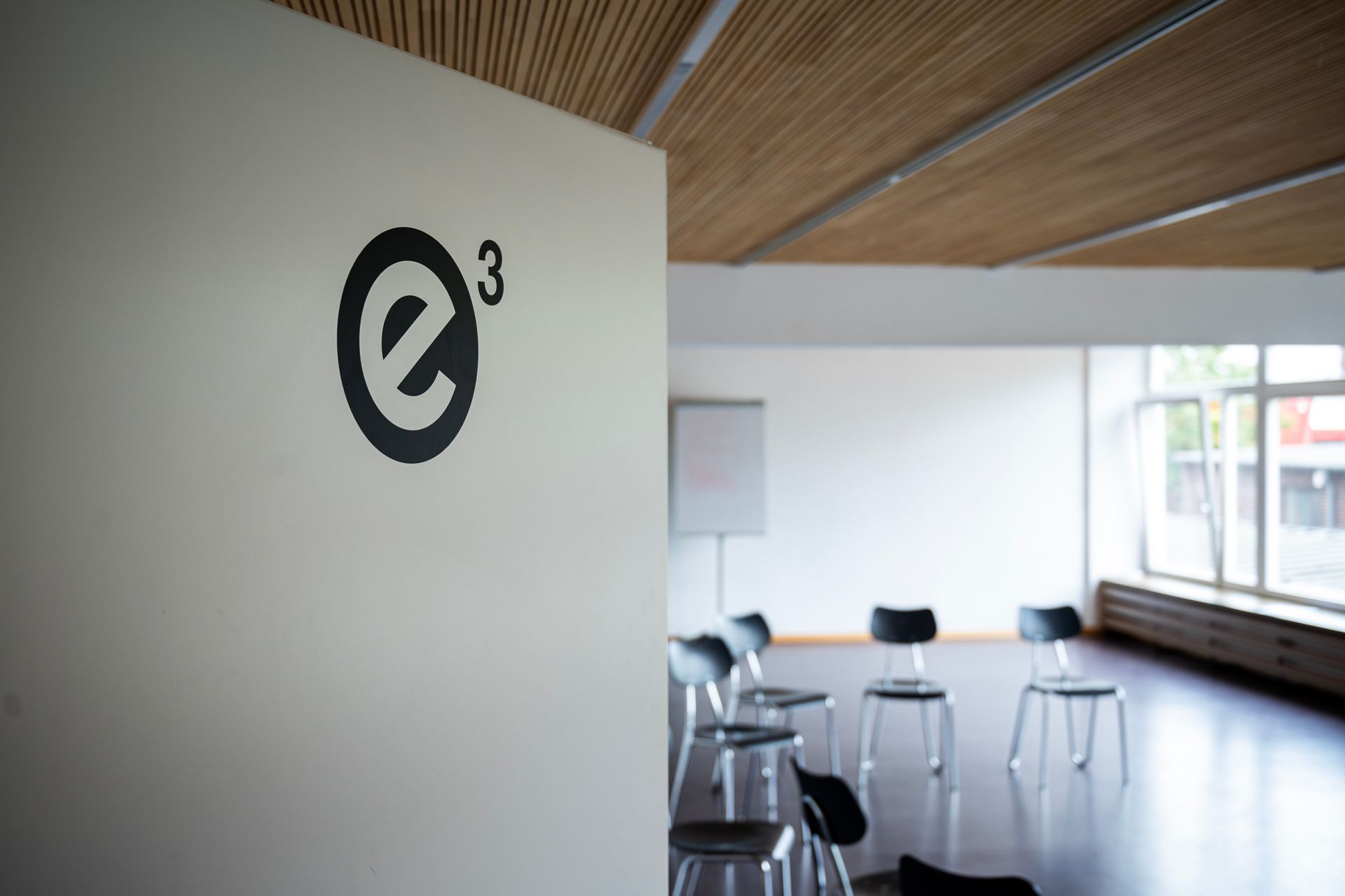 Schwarzes „e³“-Logo an einer weißen Wand in einem Raum mit Stühlen und einem Fenster.