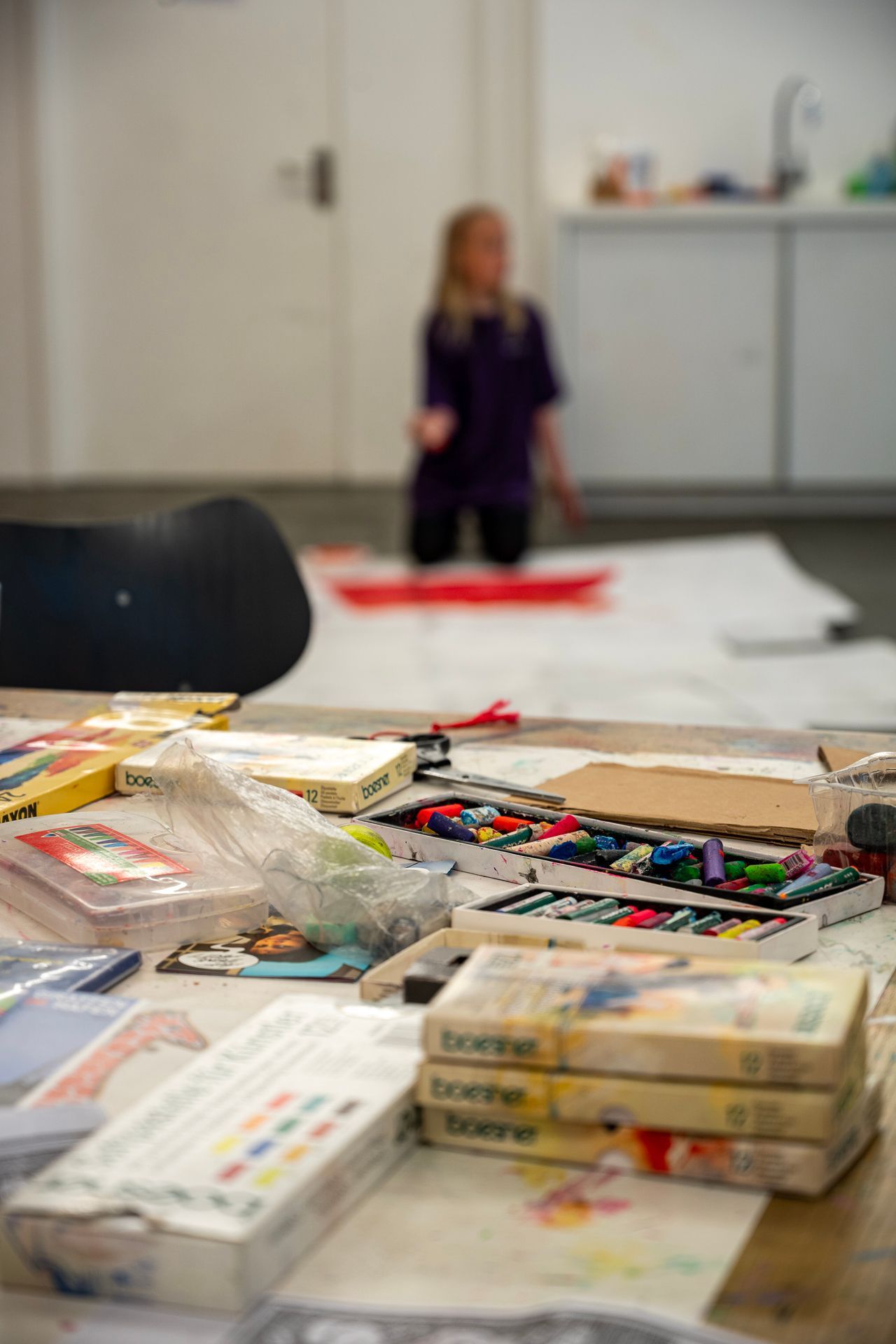 Malutensilien auf einem Tisch; im Hintergrund kniet ein Kind, das möglicherweise auf einem großen Blatt Papier zeichnet.