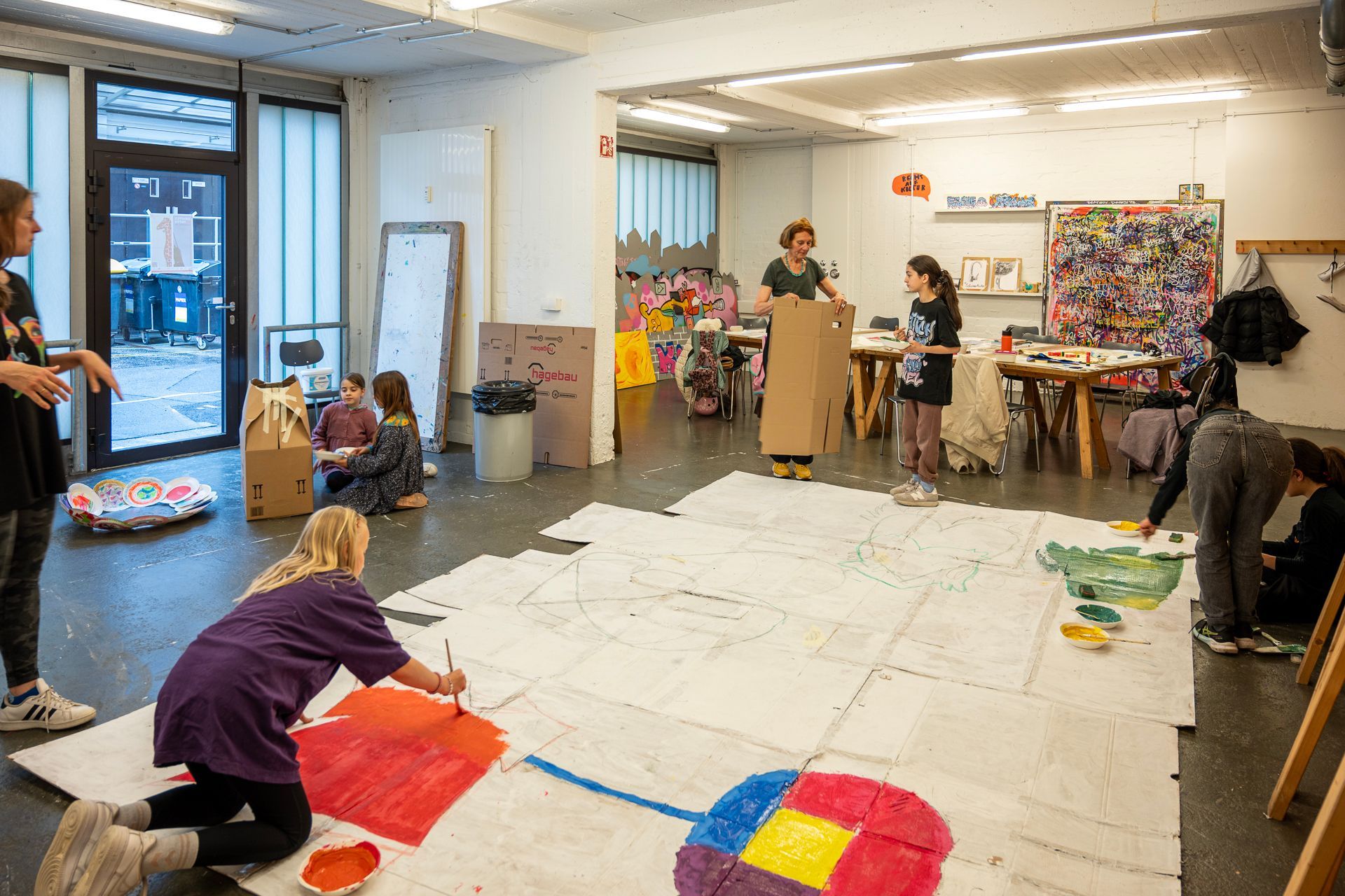 Menschen malen in einem Künstleratelier ein großes Bodenbild. Verstreute Malutensilien, Karton und fertige Kunstwerke.