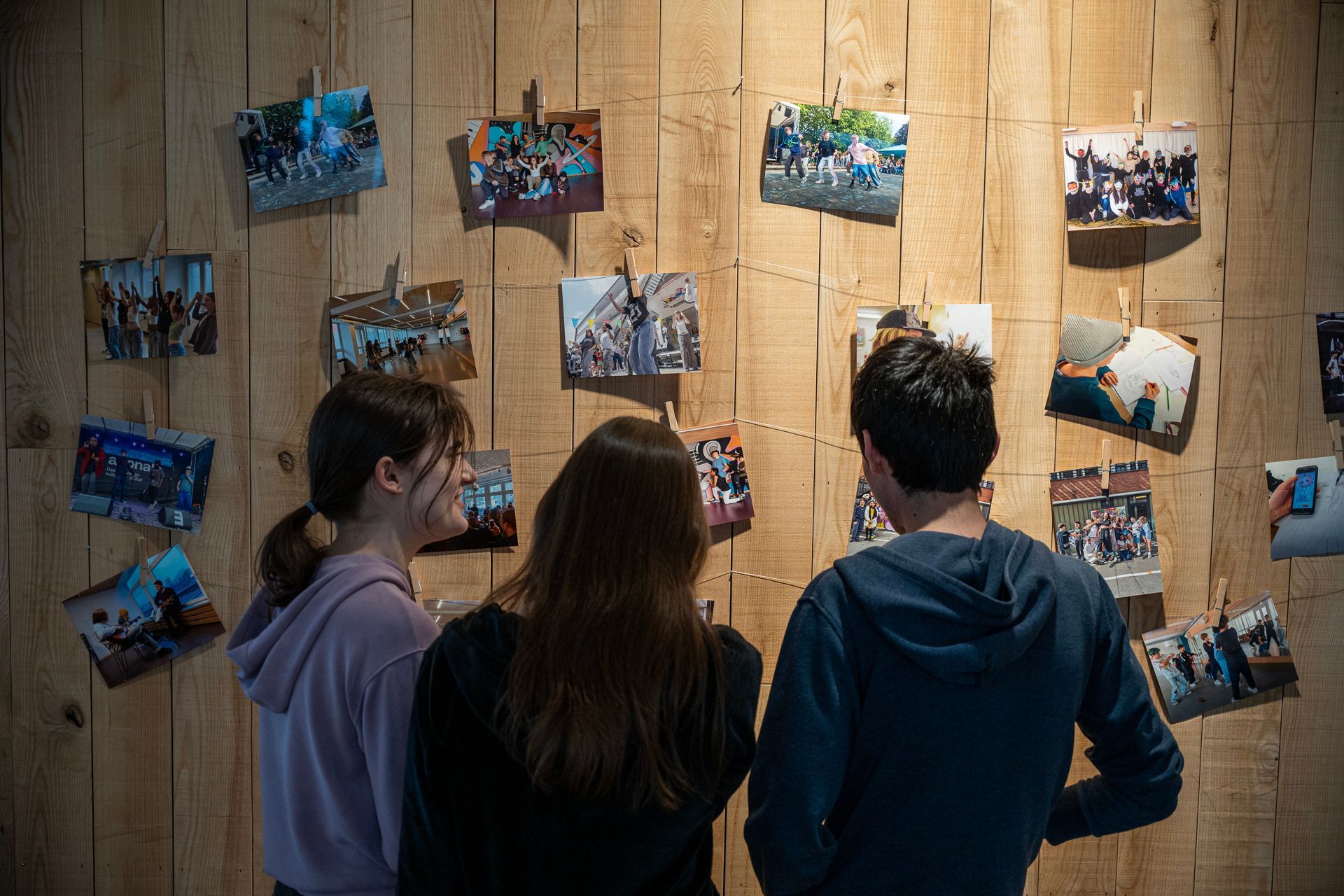 Drei Personen betrachten Fotos, die an einer Holzwand befestigt sind. Die Fotos sind mit Schnüren verbunden.