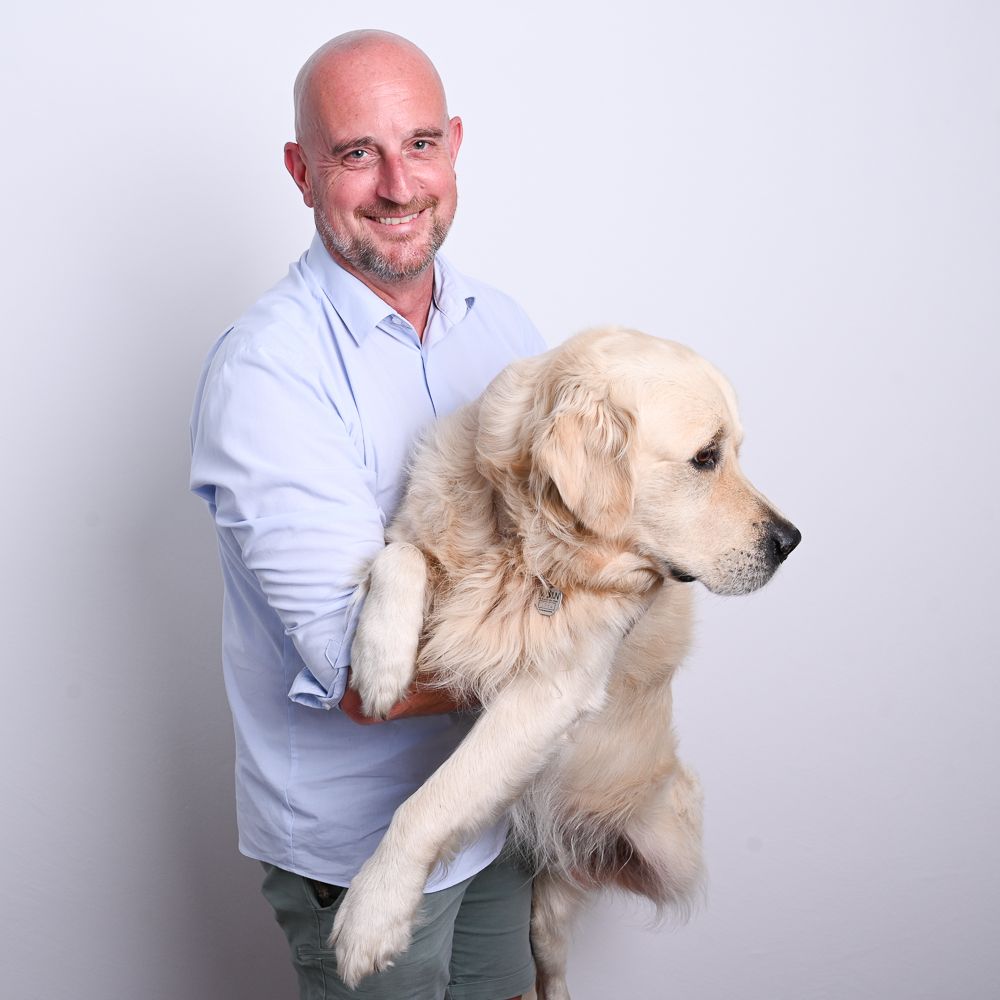 Mann hält einen Golden Retriever. Beide lächeln vor weißem Hintergrund.