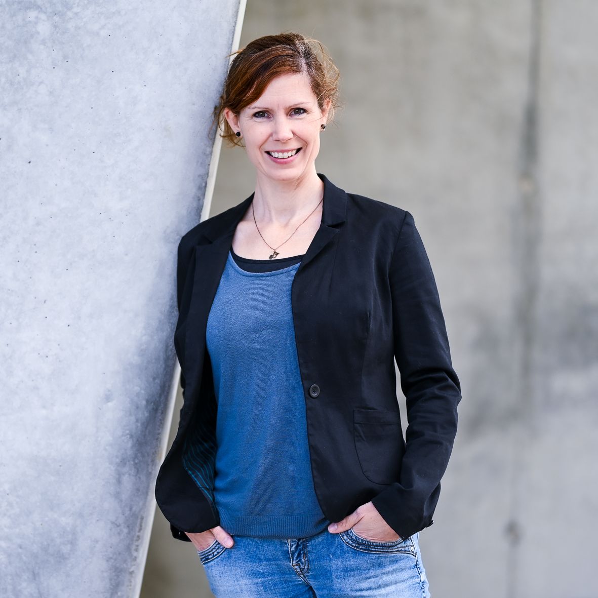 Frau in Blazer und Jeans lehnt lächelnd an einer Betonwand. Kirstin Hammerstein, hammerstein-pictures