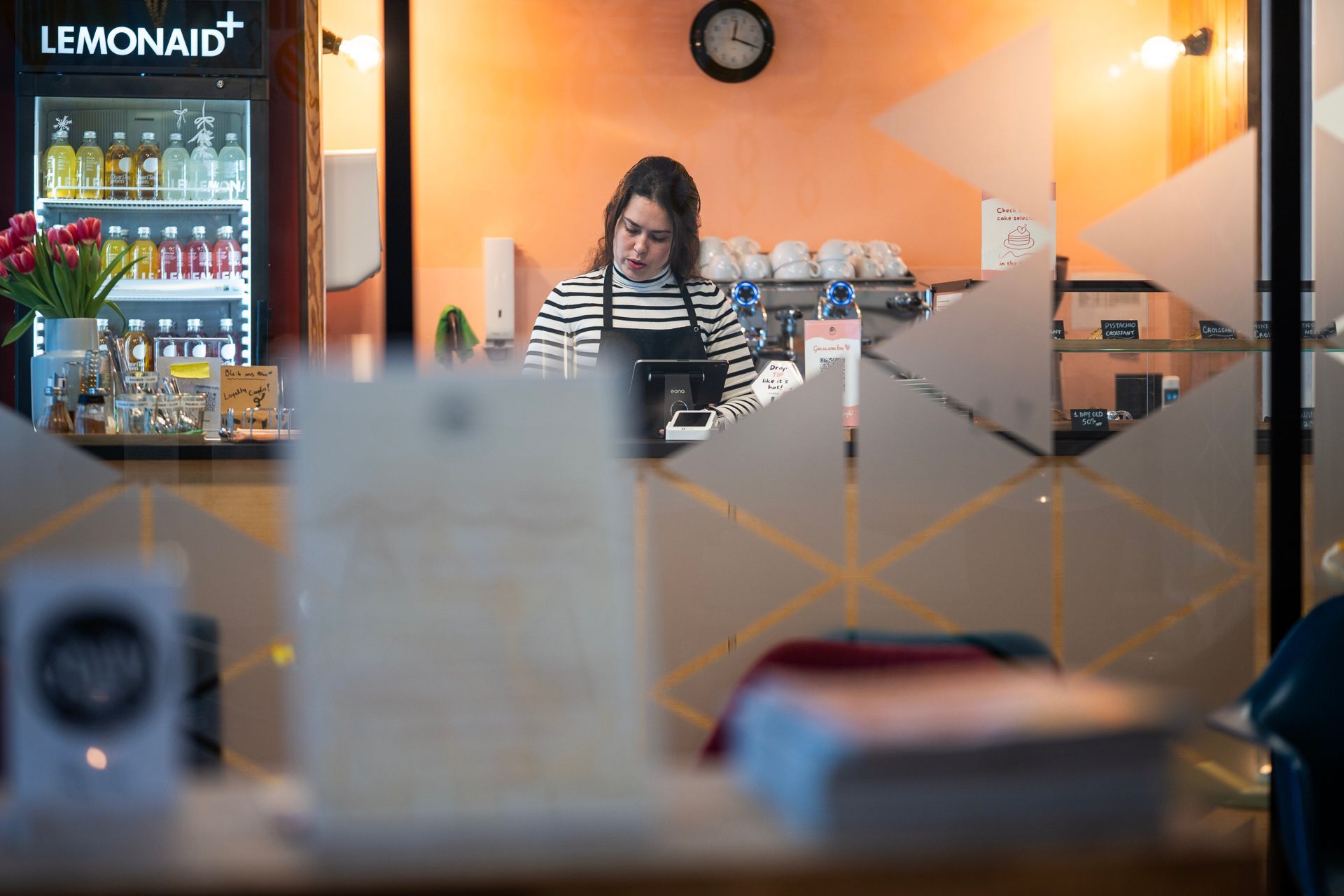 Frau in gestreiftem Hemd arbeitet hinter einem Café-Tresen, gesehen durch ein Milchglasfenster.