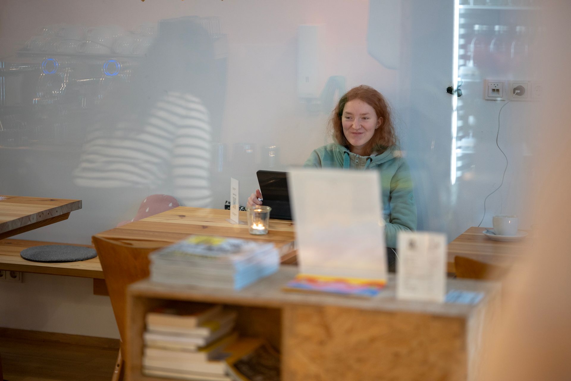 Eine rothaarige Frau sitzt an einem Holztisch in einem Café und schaut auf ein Tablet. Im Vordergrund sind Bücher und eine Kerze zu sehen.