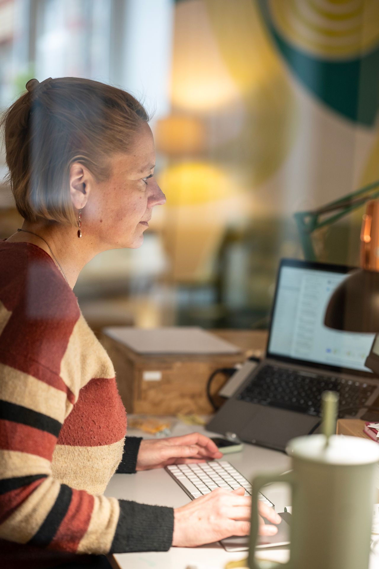 Eine Frau arbeitet an einem Laptop an einem Schreibtisch mit Tastatur, Kaffeetasse und Lampe.
