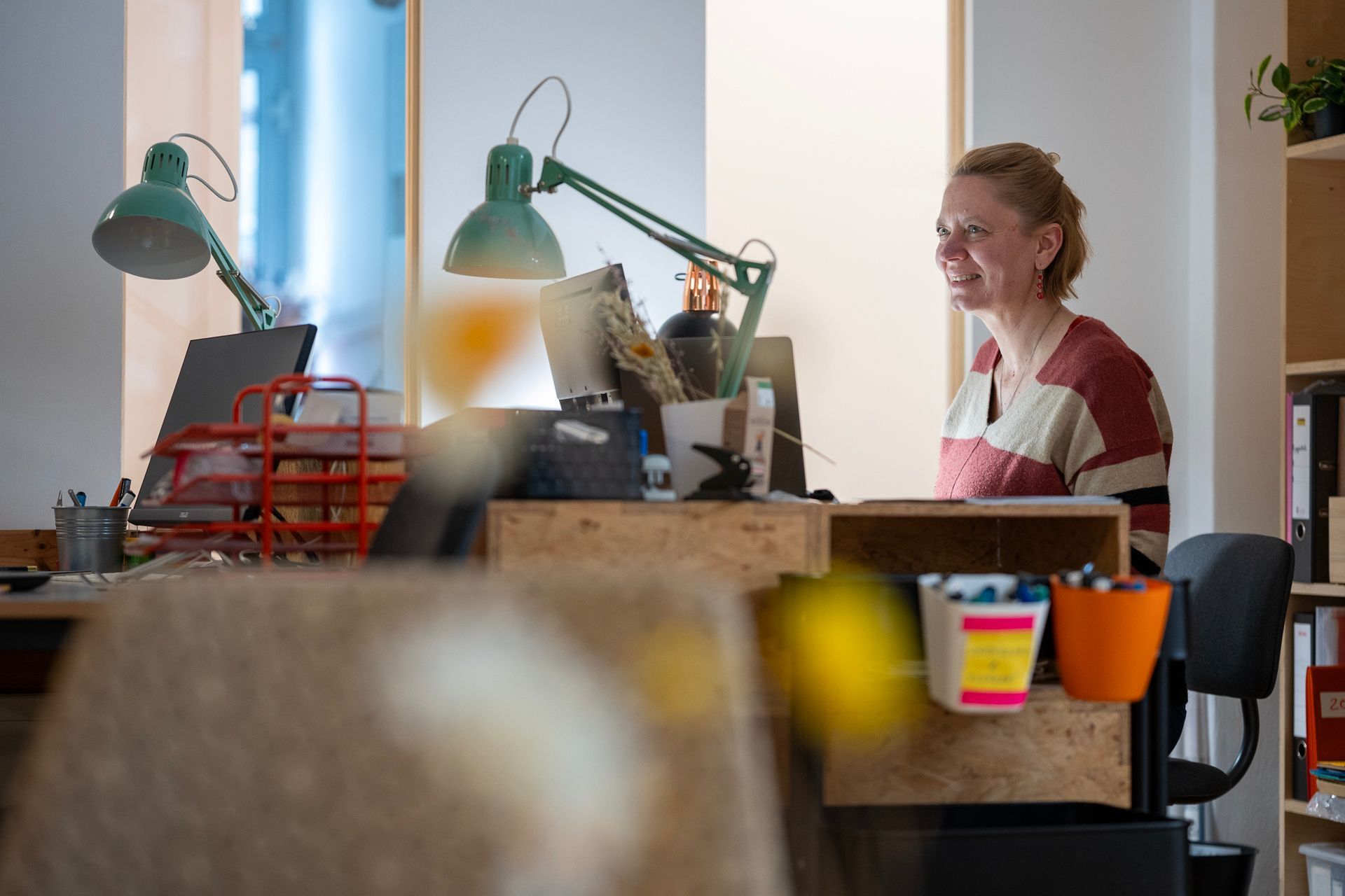 Eine Frau lächelt, während sie an einem vollgestellten Schreibtisch mit zwei grünen Lampen in einem hell erleuchteten Büro sitzt.