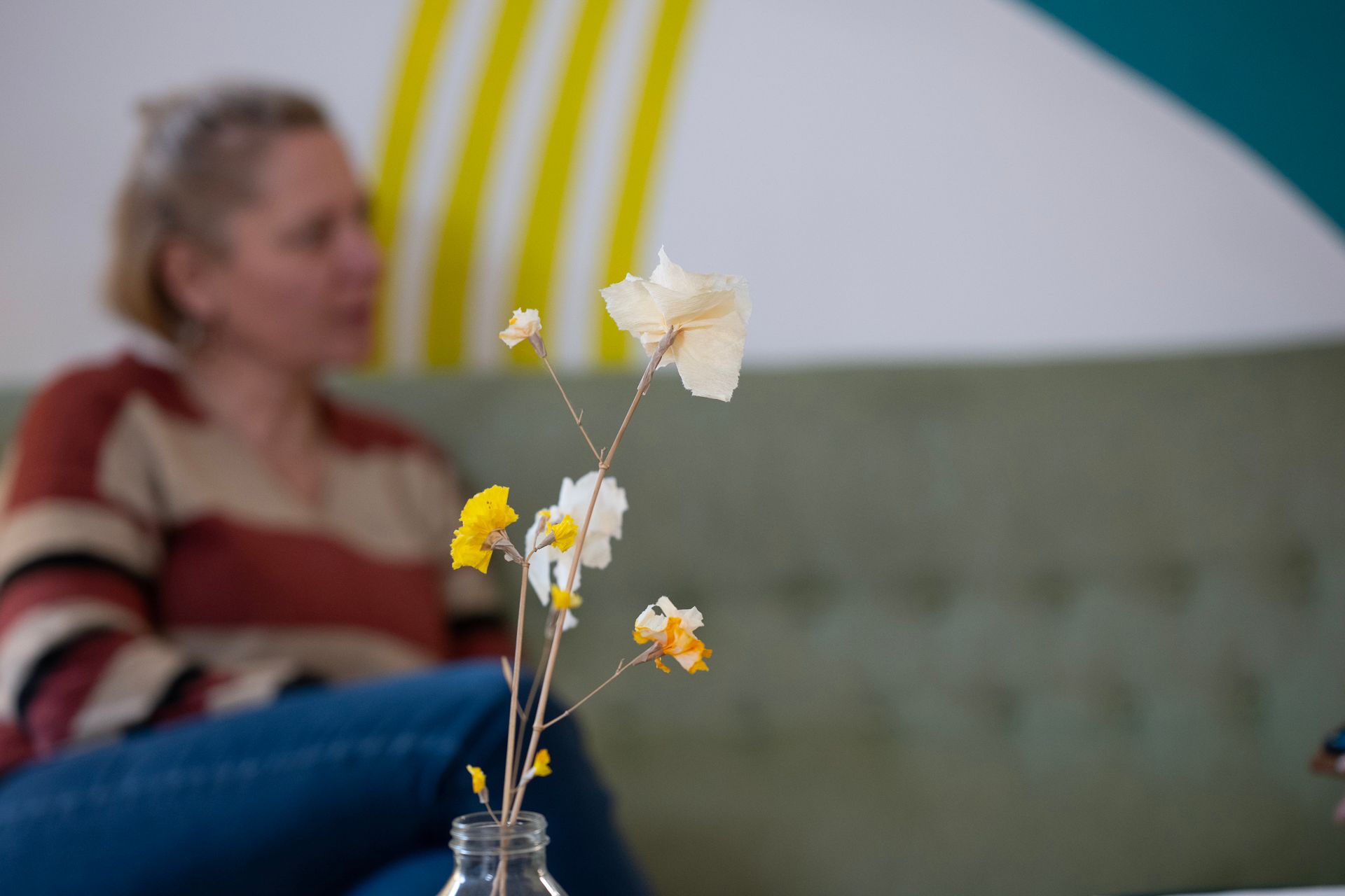 Nahaufnahme einer Vase mit Blumen; im Hintergrund eine Frau auf einem grünen Sofa. Helle Wandgestaltung im Hintergrund.