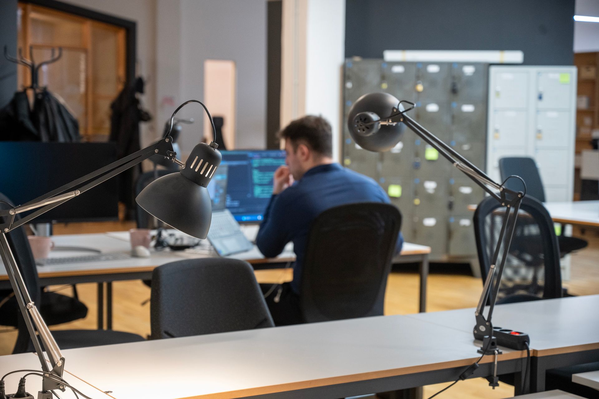 Eine Person arbeitet an einem Schreibtisch in einem Büro. Zwei Lampen beleuchten den Arbeitsbereich.