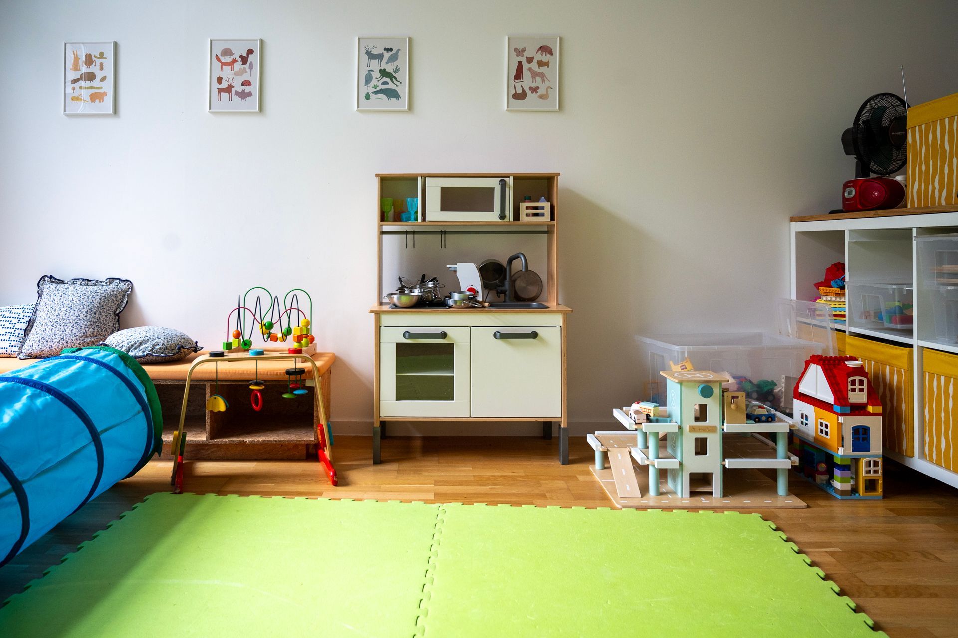 Spielzimmer mit Spielküche, Holzspielzeug, Spieltunnel und Stauraum. Grüne Spielmatte auf dem Boden.