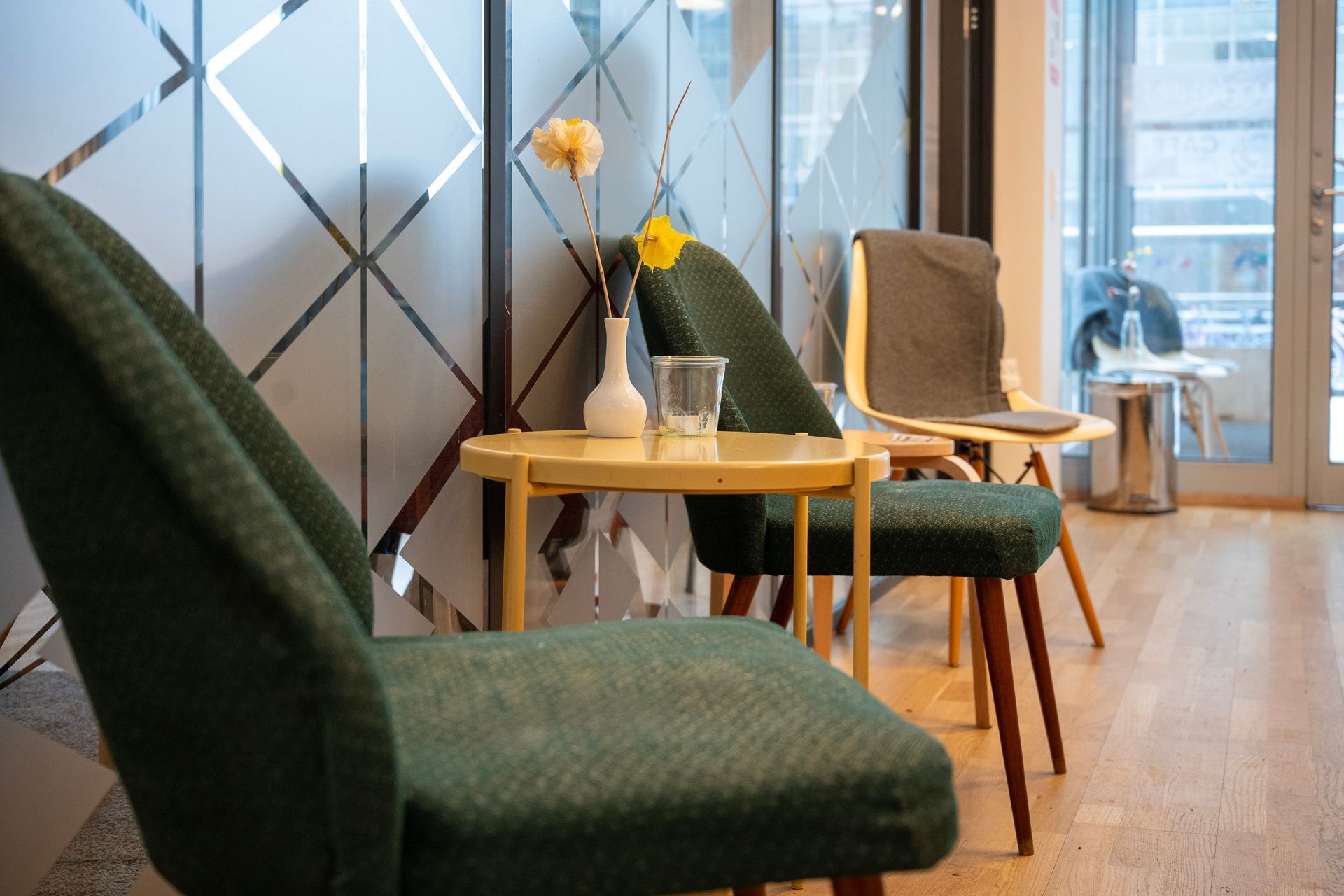Grüne und graue Stühle flankieren einen kleinen Tisch mit einer Vase und einem Glas vor einer gemusterten Glaswand.