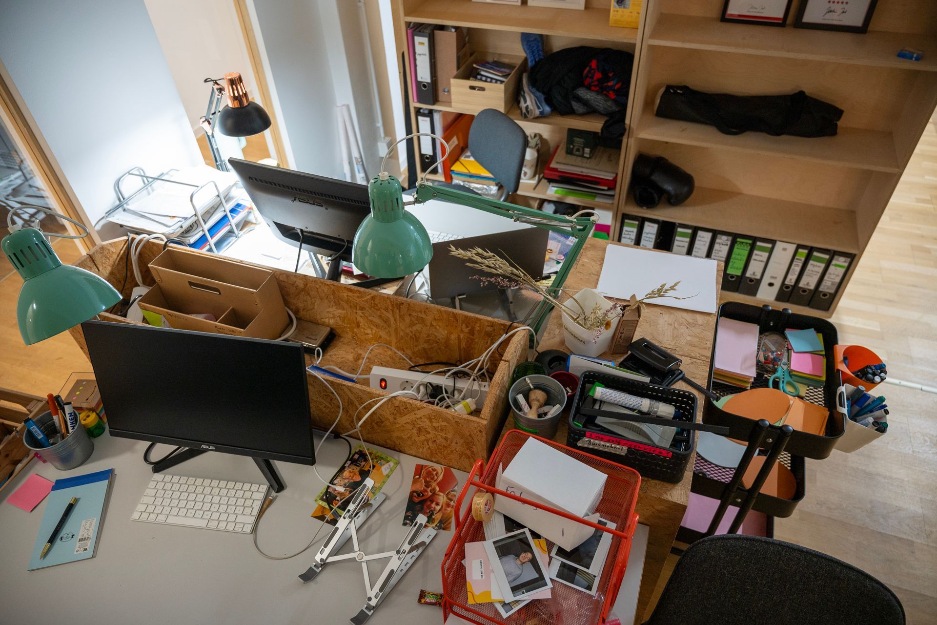 Ein vollgestellter Schreibtisch mit Computermonitor, Lampen, Papieren und verschiedenen Büroartikeln.