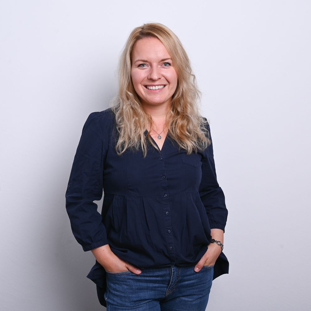 Frau mit blonden Haaren, lächelnd, trägt eine dunkelblaue Bluse und Jeans, die Hände in den Taschen.