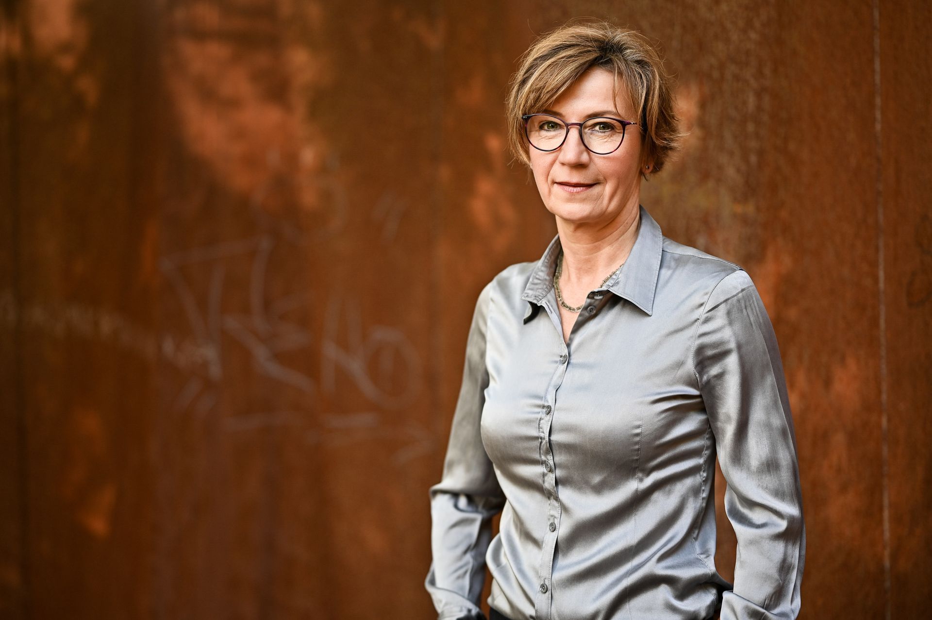 Frau mit Brille, silberner Bluse, Hände in den Taschen, steht vor rostiger Metallwand. Kirstin Hammerstein, hammerstein-pictures