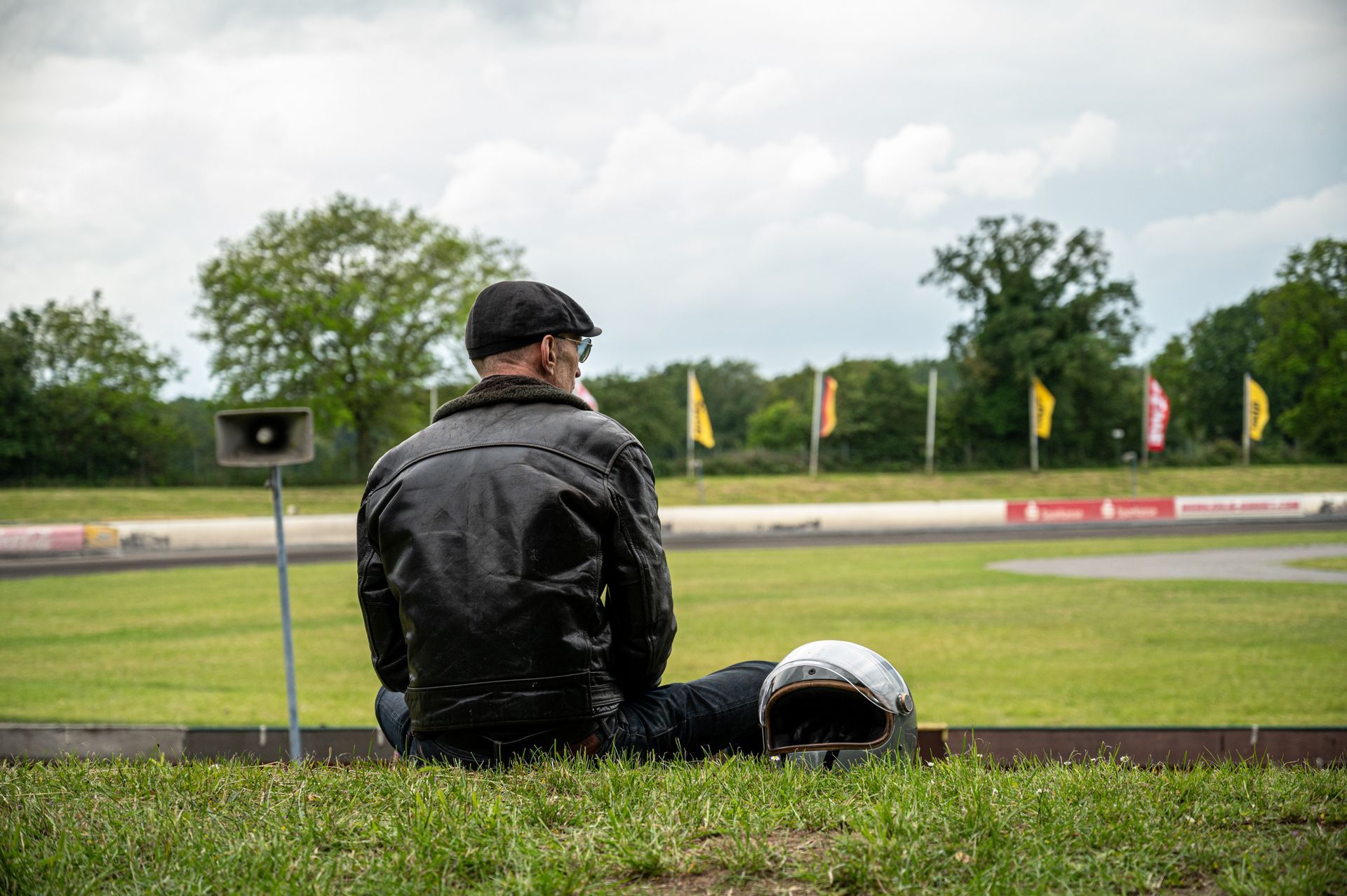 Mann in Lederjacke und Mütze sitzt auf Gras, Helm neben ihm, und blickt auf eine Rennstrecke. Kirstin Hammerstein, hammerstein-pictures