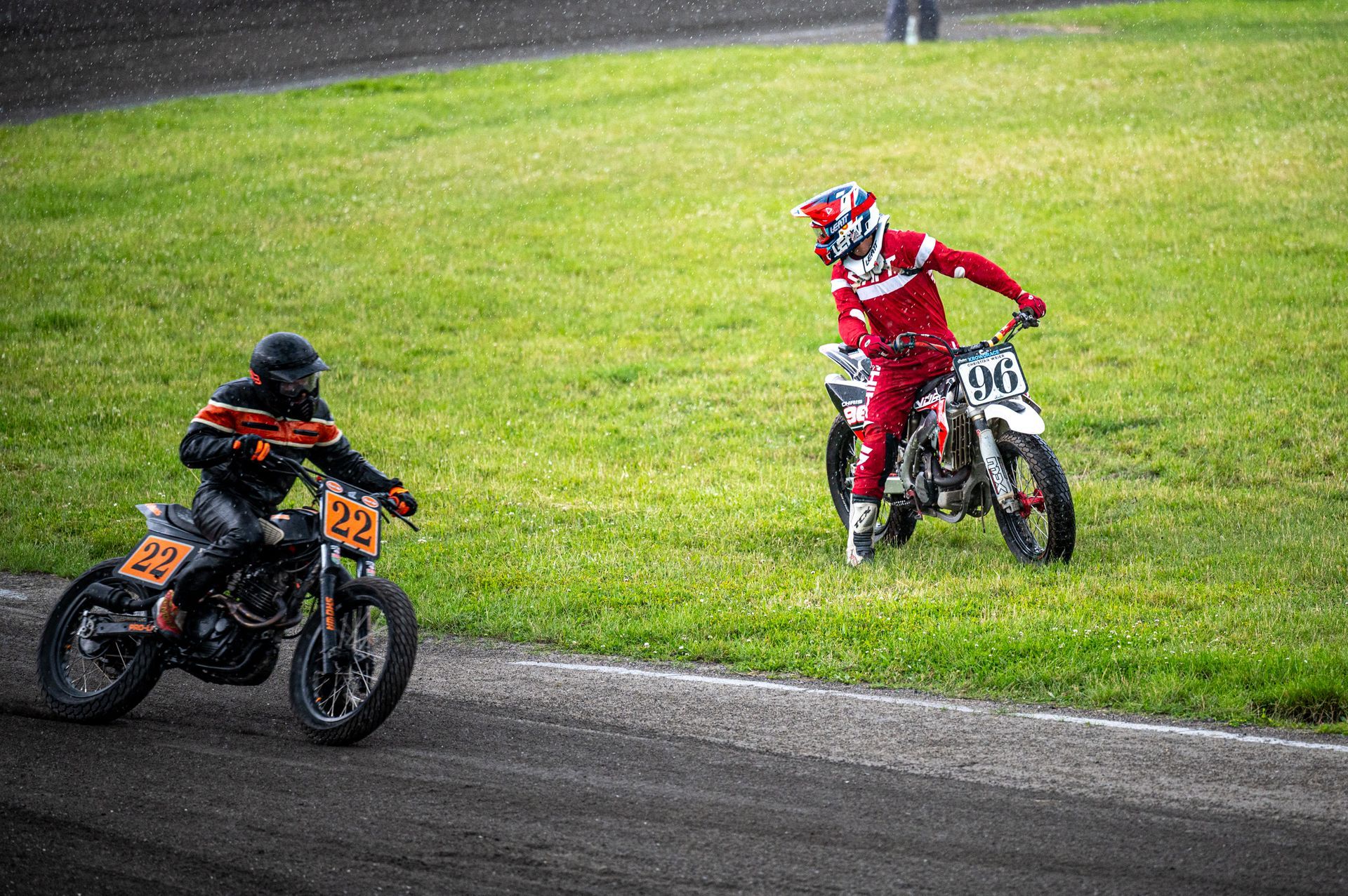 Zwei Flat-Track-Motorradrennfahrer auf einer Grasstrecke. Einer in roter Ausrüstung, einer in orangefarbener. Kirstin Hammerstein, hammerstein-pictures