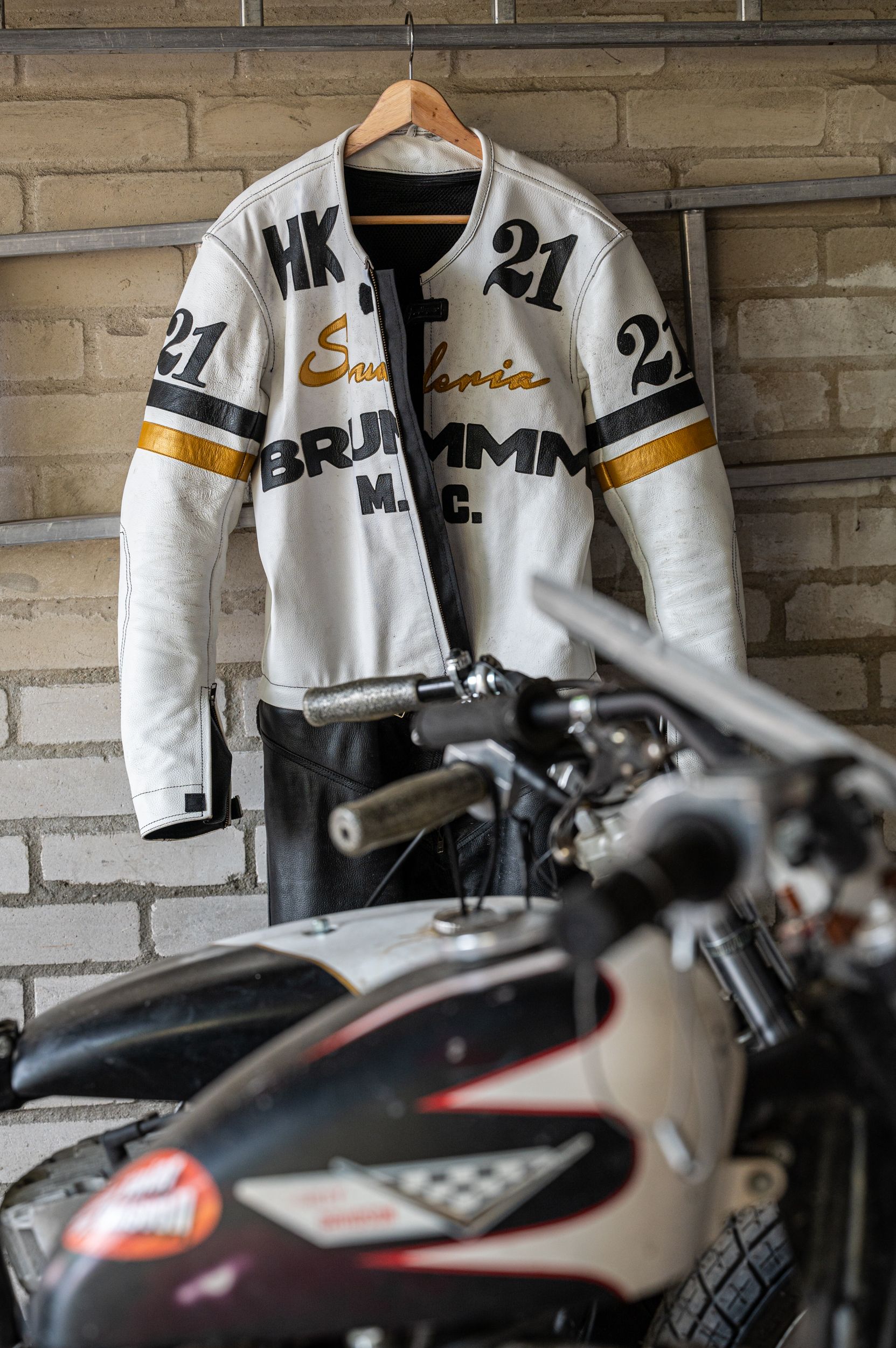 Weiße Motorradjacke mit schwarzen und goldenen Akzenten, die auf einem Kleiderbügel neben einem Motorrad hängt. Kirstin Hammerstein, hammerstein-pictures