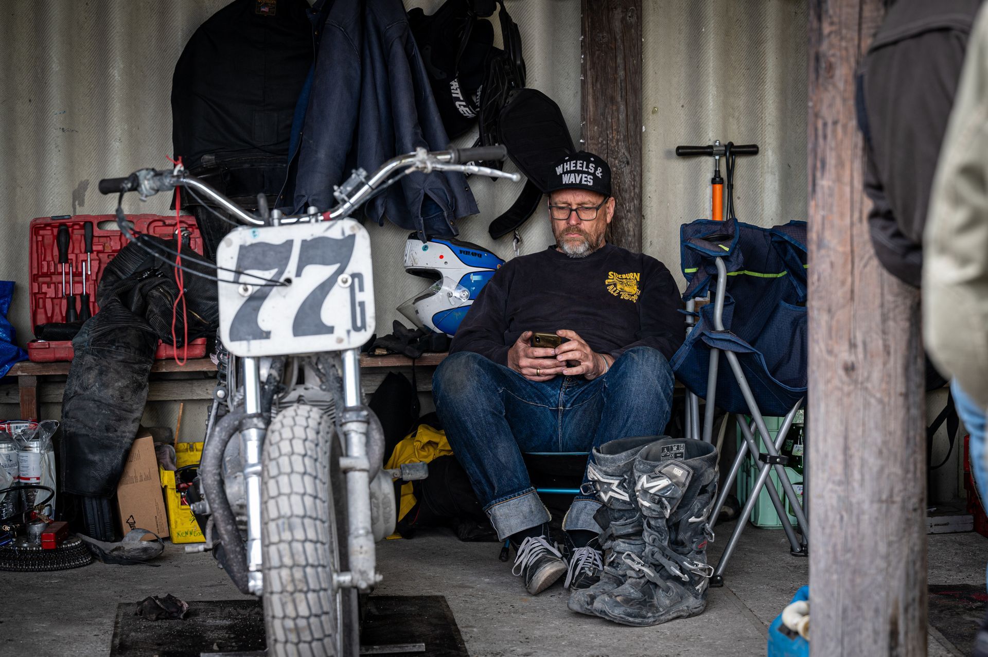 Mann mit Mütze und Jeans sitzt neben einem Dirt-Track-Motorrad und schaut in einer Garage auf sein Telefon. Kirstin Hammerstein, hammerstein-pictures