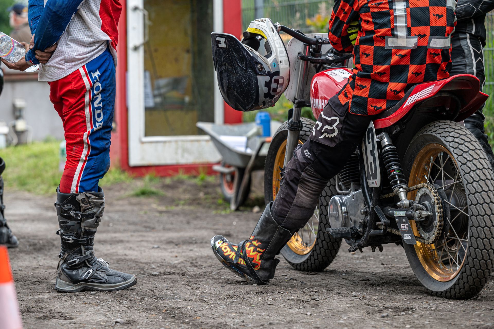 Zwei Motorradrennfahrer bereiten sich vor. Einer steht neben dem Motorrad, der andere sitzt. Beide tragen Schutzkleidung; die Kulisse scheint eine Rennstrecke zu sein. Kirstin Hammerstein, hammerstein-pictures