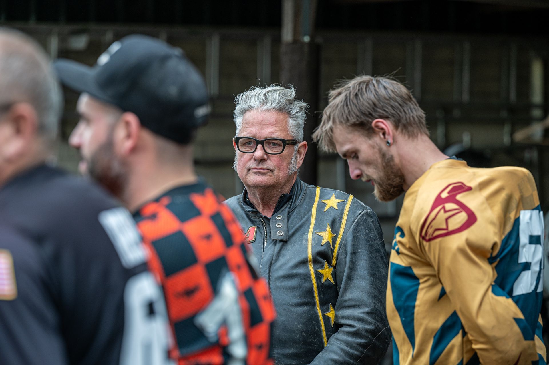 Ein Mann in einer Lederjacke mit Sternen sieht einen Mann in einem goldenen Rennanzug an, in dessen Nähe sich andere Männer befinden. Kirstin Hammerstein, hammerstein-pictures