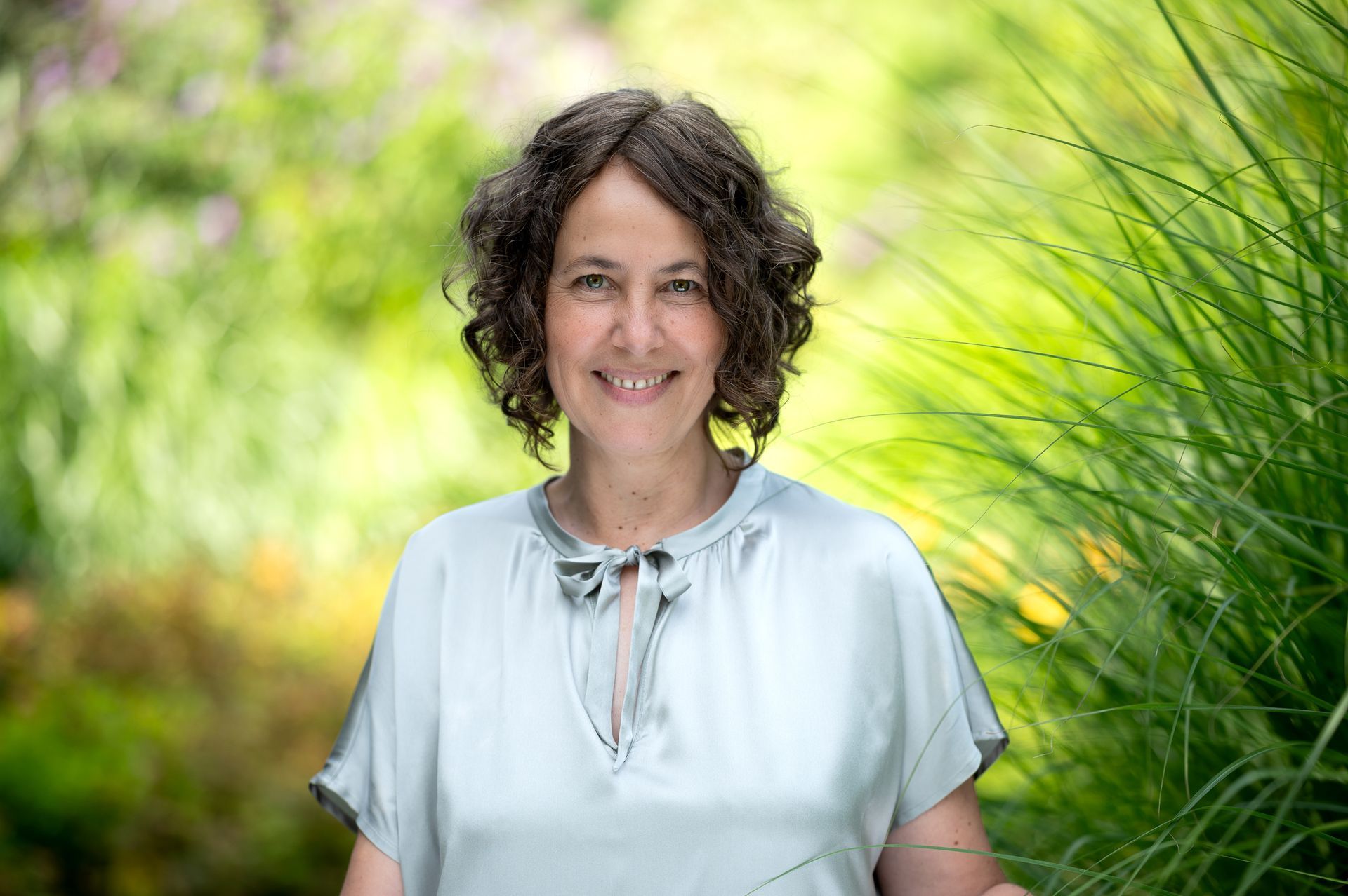 Frau mit lockigem braunem Haar lächelt in einem Garten und trägt eine hellblaue Bluse. Kirstin Hammerstein, hammerstein-pictures