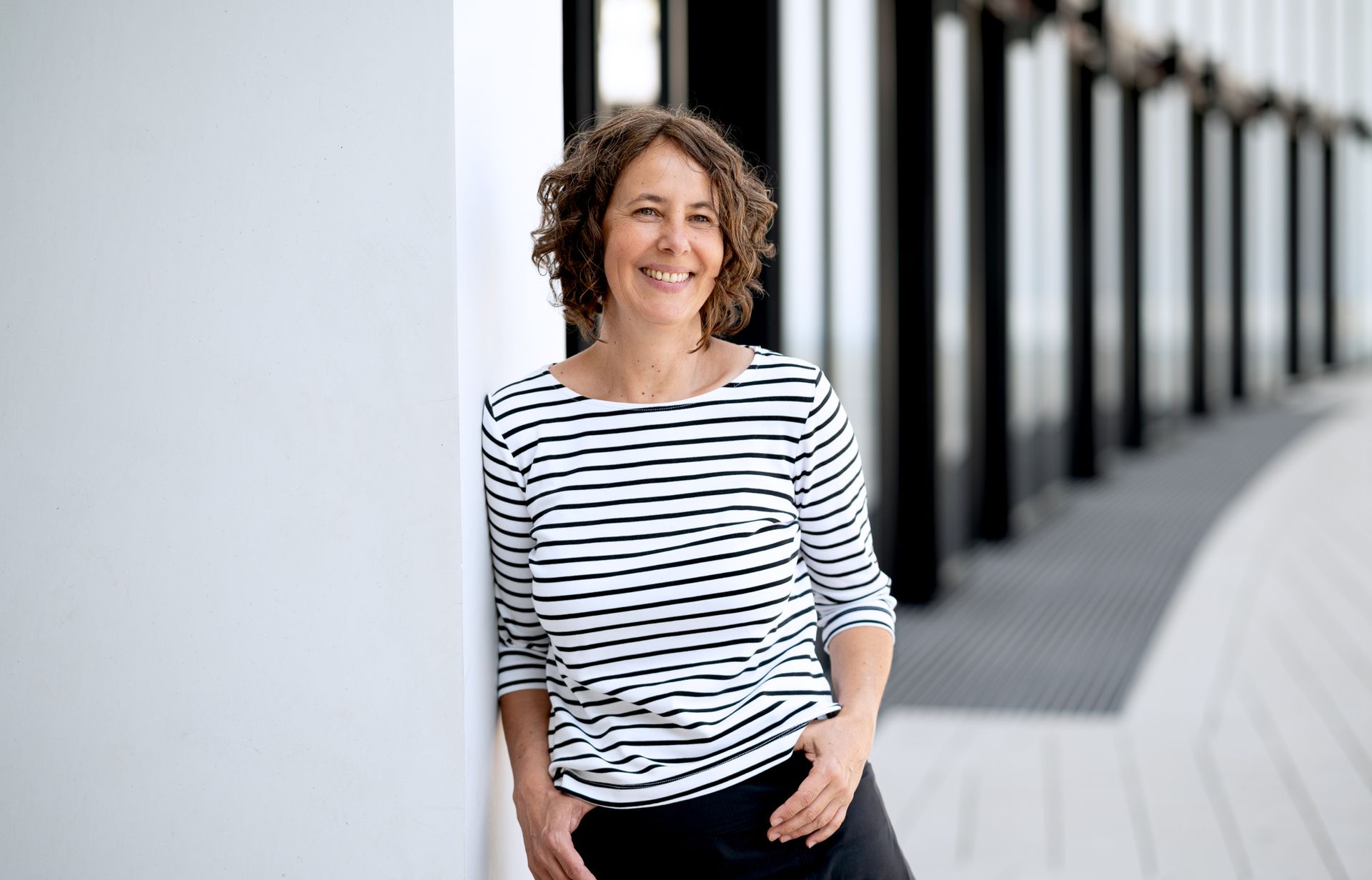 Frau mit lockigem Haar in gestreiftem Hemd lächelt, lehnt an weißer Wand, Hände in den Taschen. Kirstin Hammerstein, hammerstein-pictures