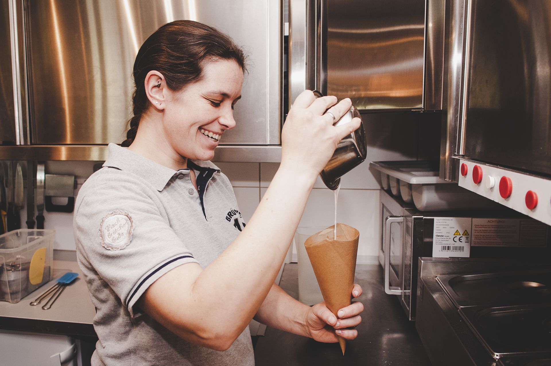 Kirstin Hammerstein, hammerstein-pictures, Eine Frau gießt in einer Großküche Flüssigkeit aus einem Behälter in eine Papiertüte.