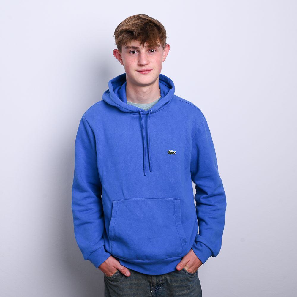 Ein Teenager in einem blauen Kapuzenpulli, die Hände in den Taschen, lächelt vor einem schlichten Hintergrund in die Kamera.