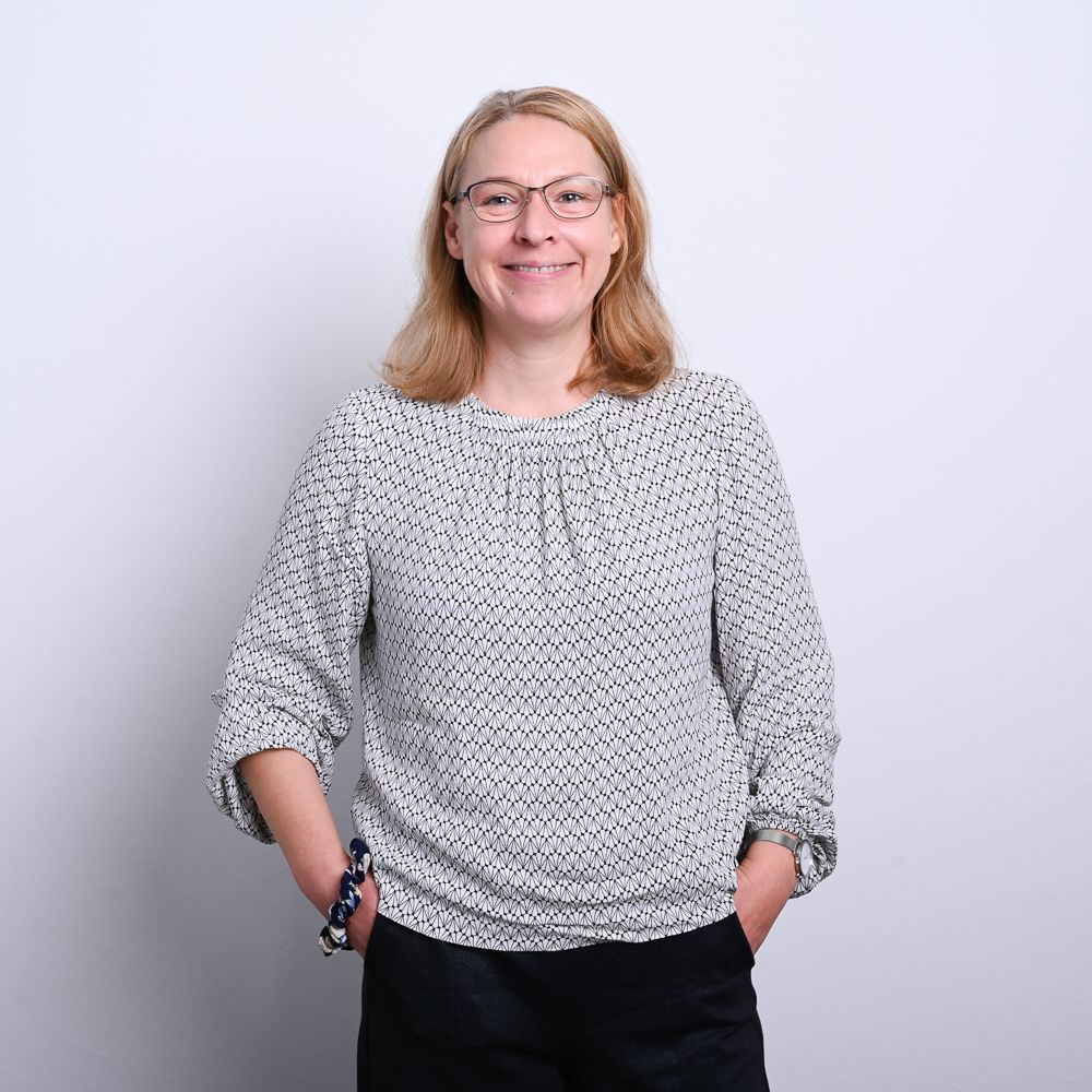 Frau mit Brille und gemustertem Oberteil lächelt, Hände in den Taschen.