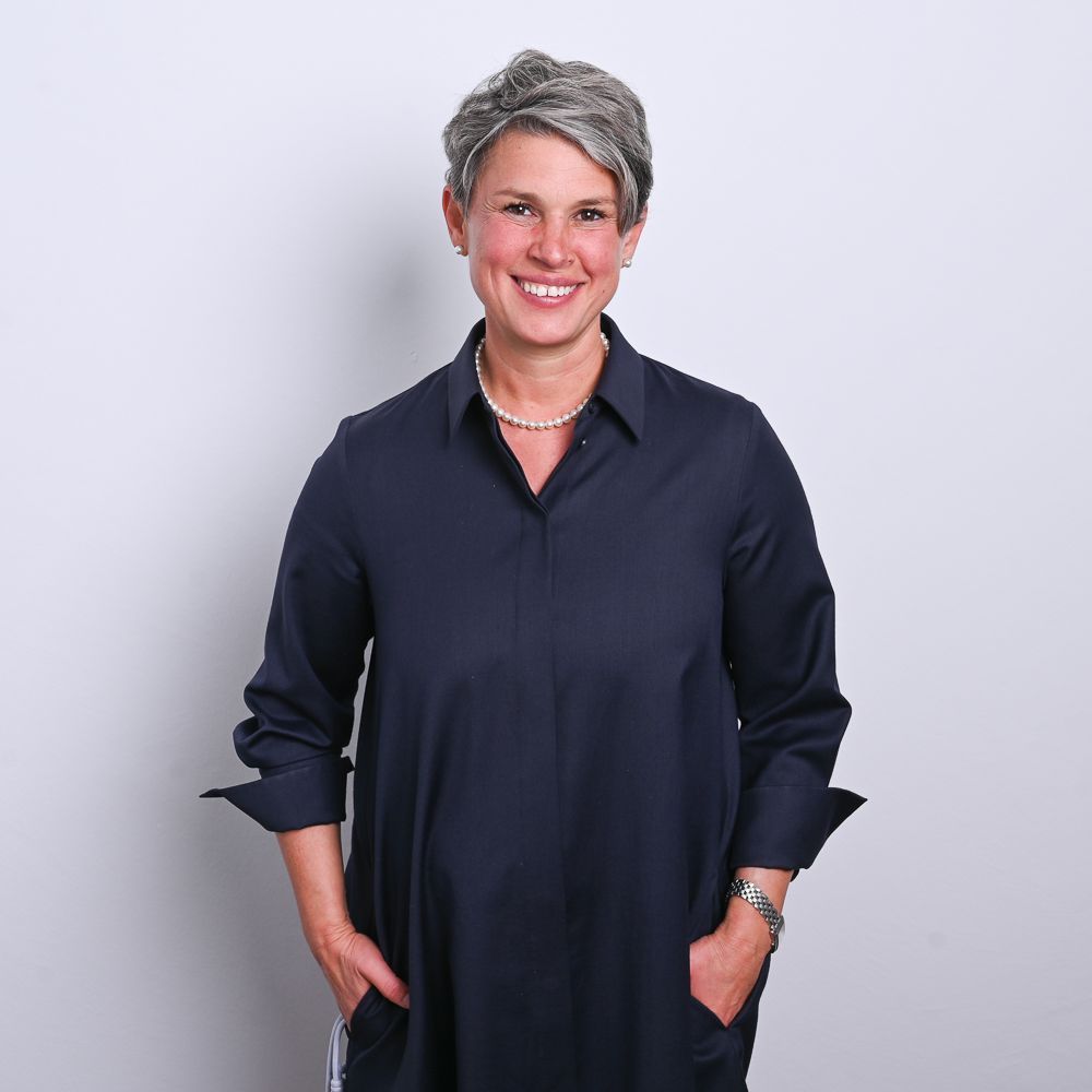 Eine Frau mit kurzem, grauem Haar lächelt, die Hände in den Taschen eines marineblauen Kleides. Vor weißem Hintergrund.