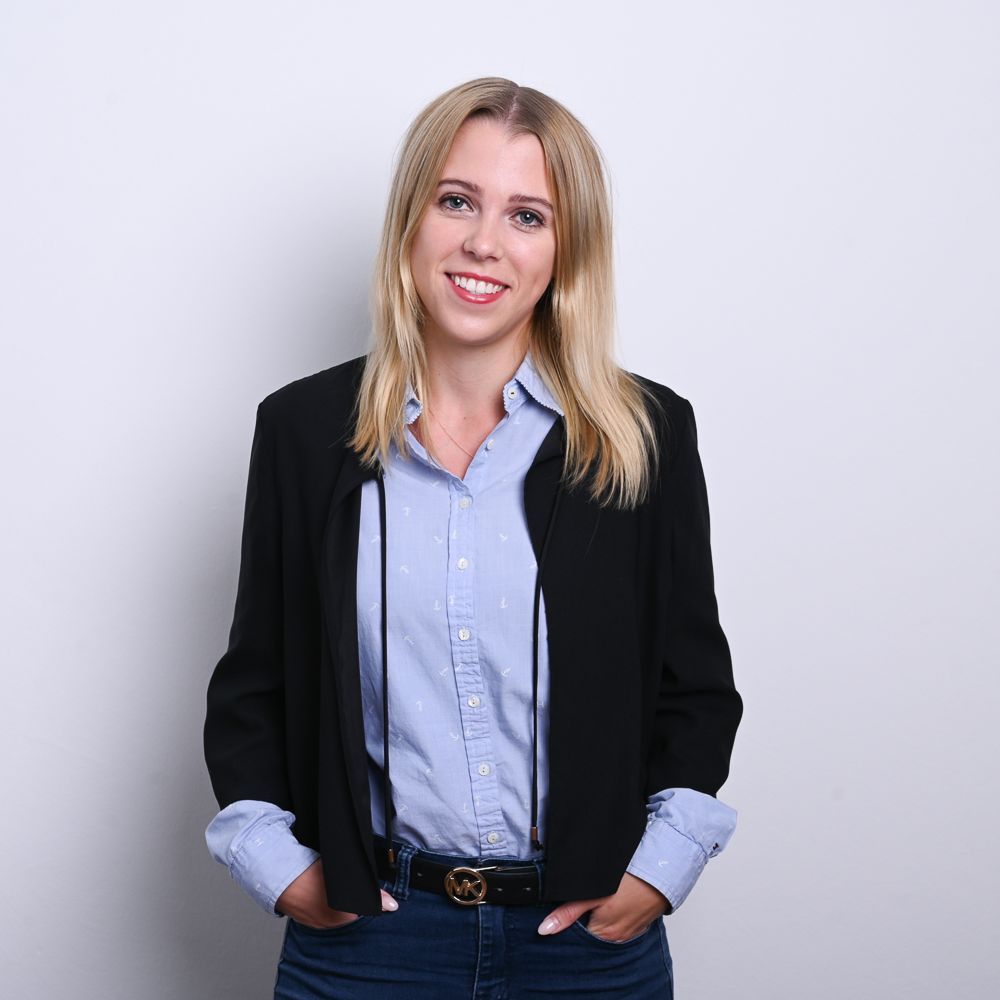 Eine Frau in Blazer und Hemd mit Knöpfen lächelt, die Hände in den Taschen, vor einem weißen Hintergrund.