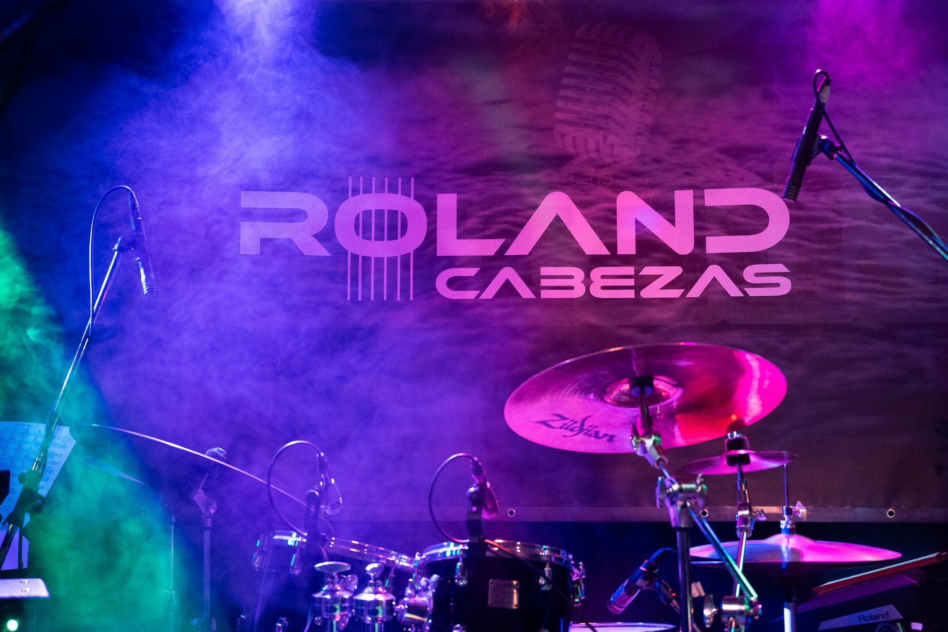 Bandaufbau auf der Bühne mit Roland Cabezas Logo, Schlagzeug, Becken, Mikrofonen und Bühnenbeleuchtung.