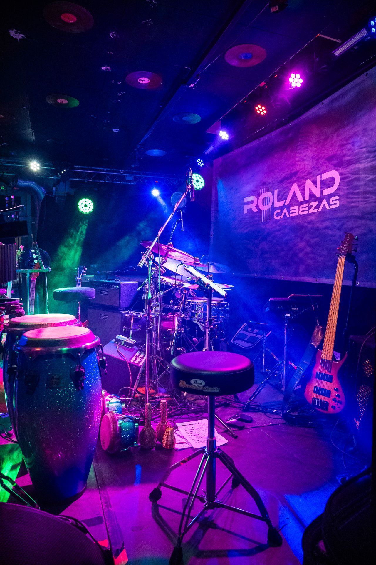 Eine in violettes und blaues Licht getauchte Bühne. Ein Schlagzeug, ein Bass und Congas sind für einen Auftritt aufgebaut.