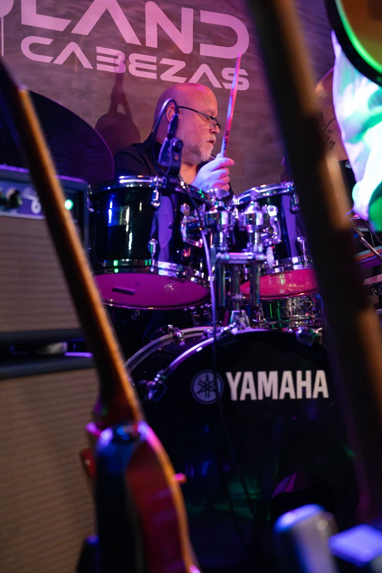 Ein Schlagzeuger spielt auf der Bühne ein Yamaha-Schlagzeug, das teilweise von Gitarrenhälsen verdeckt wird. Lila und grüne Bühnenbeleuchtung.
