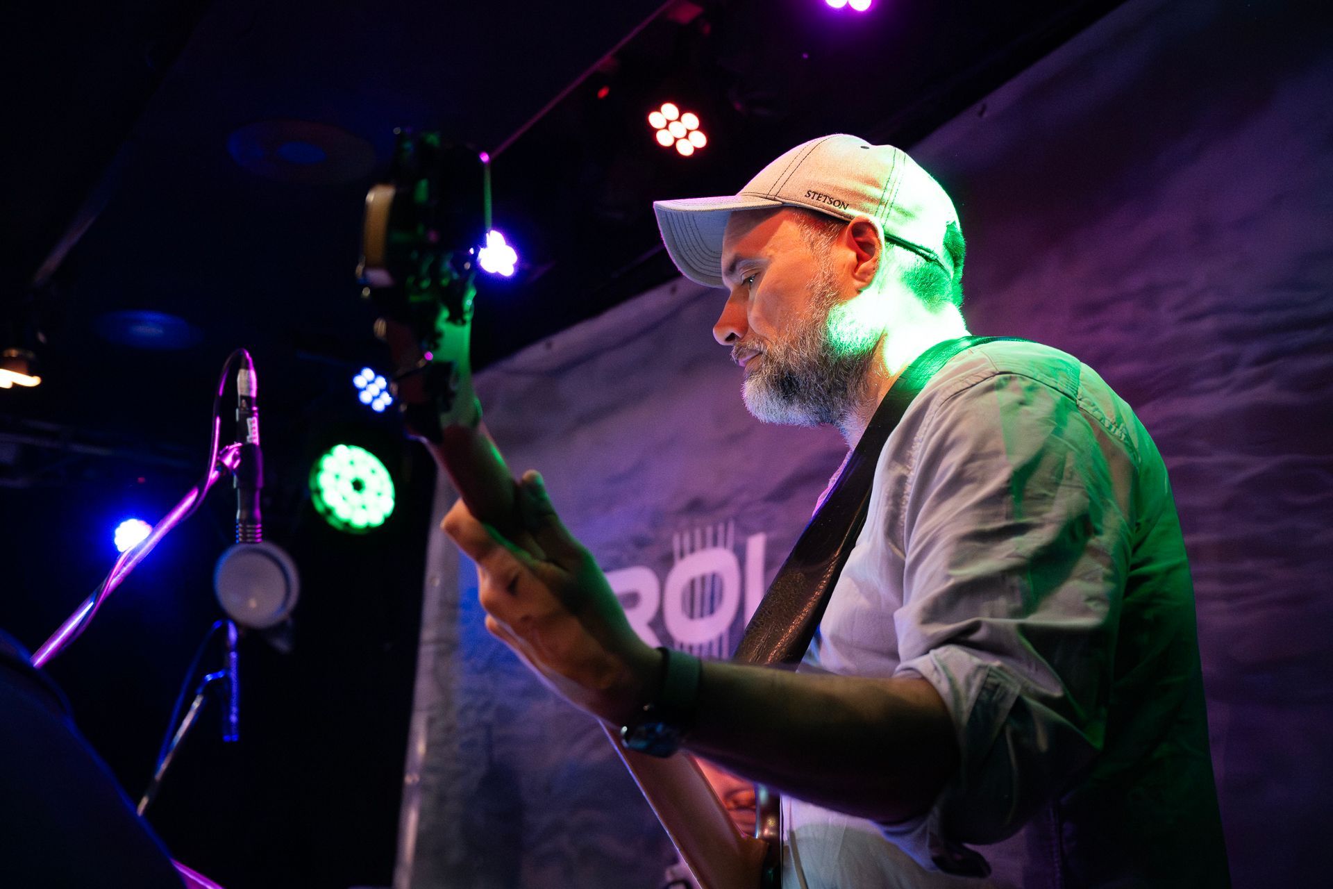 Ein Mann mit Hut spielt auf der Bühne Bassgitarre. Grüne und blaue Bühnenlichter beleuchten die Szene.