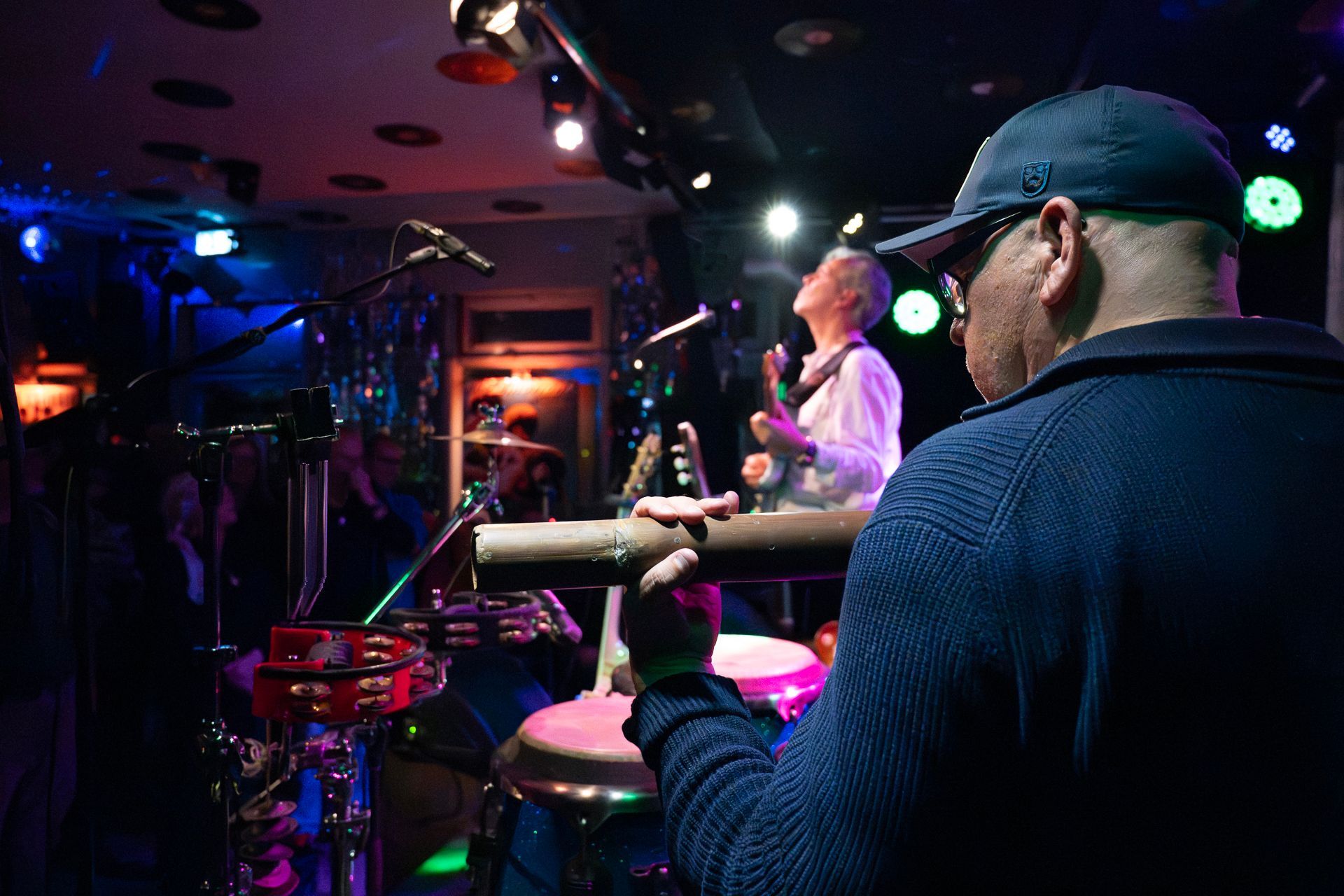 Ein Mann mit Kappe beobachtet eine Band, die in einem schwach beleuchteten Club auf der Bühne spielt.
