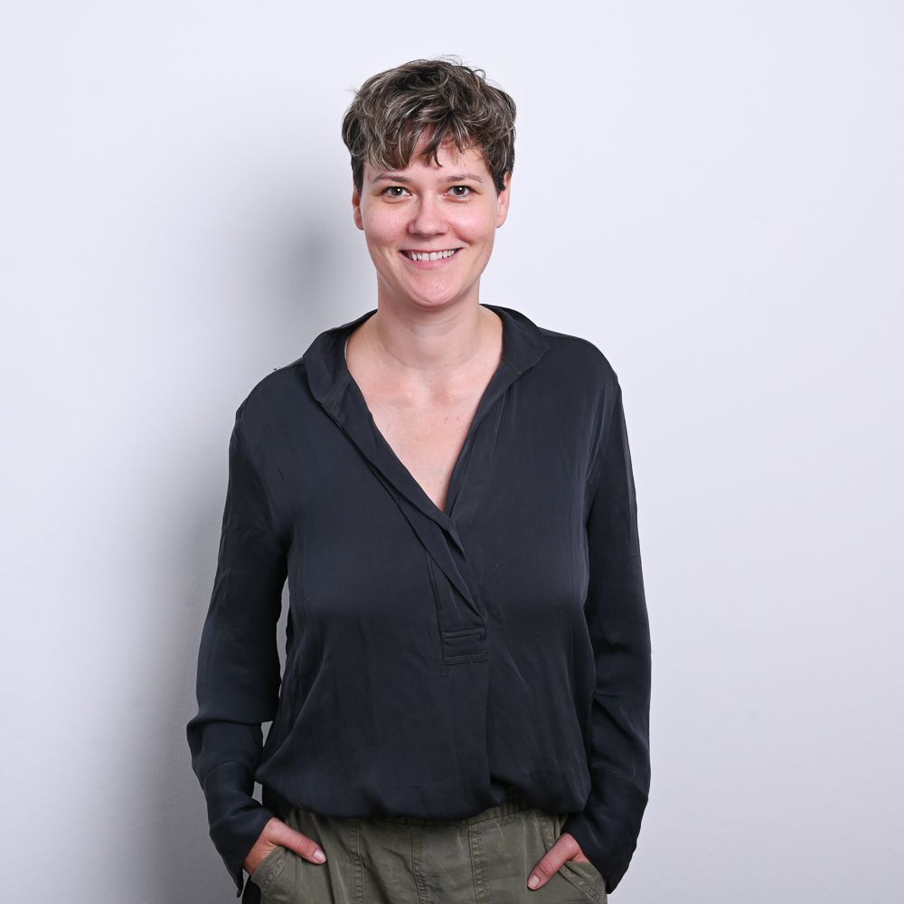 Eine Frau mit kurzen Haaren lächelt. Sie trägt ein dunkles Oberteil und eine olivgrüne Hose und hat die Hände in den Taschen.