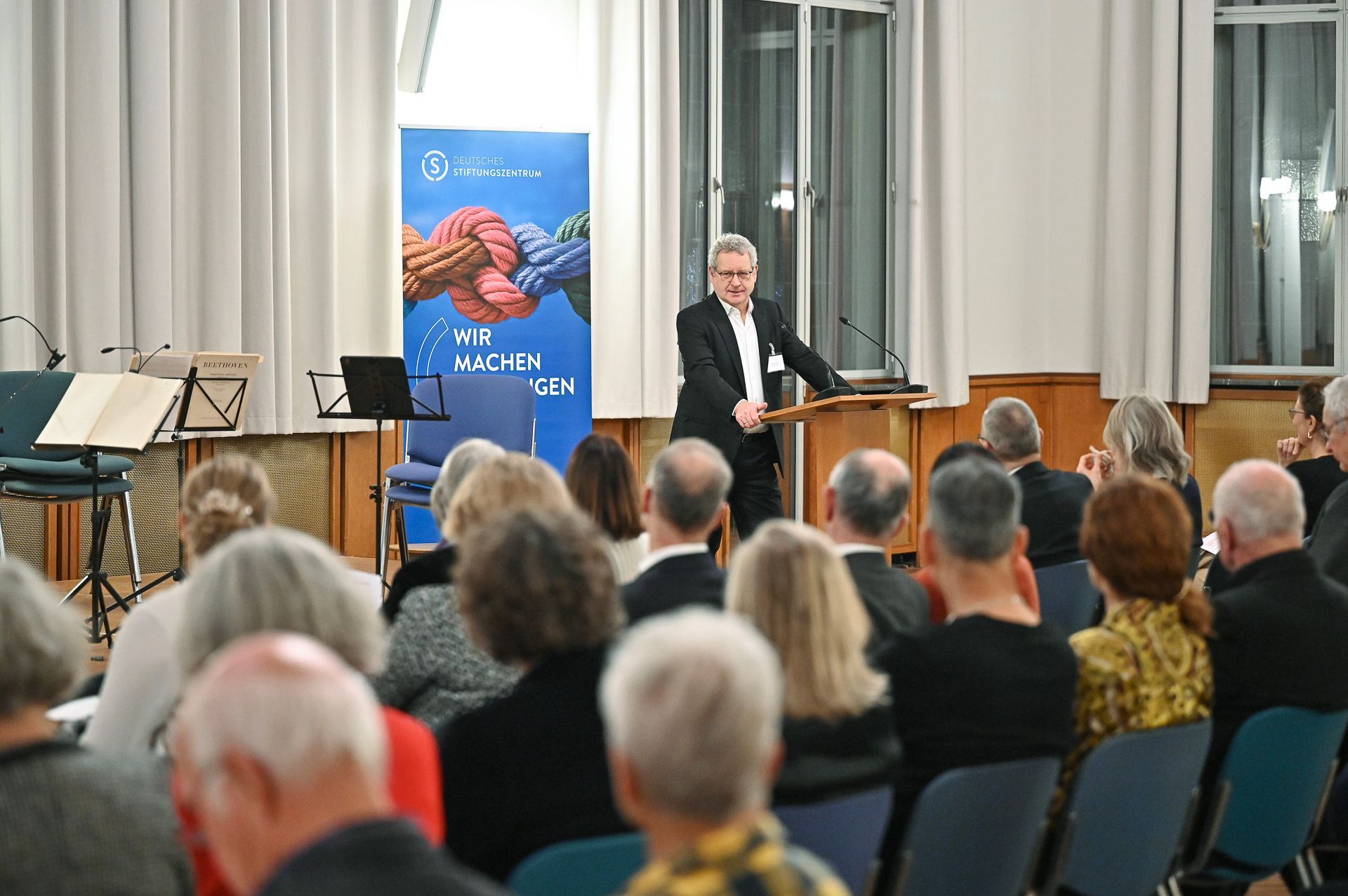 Ein Mann dirigiert ein Orchester und steht vor einem sitzenden Publikum in einem Auditorium mit einem Banner.