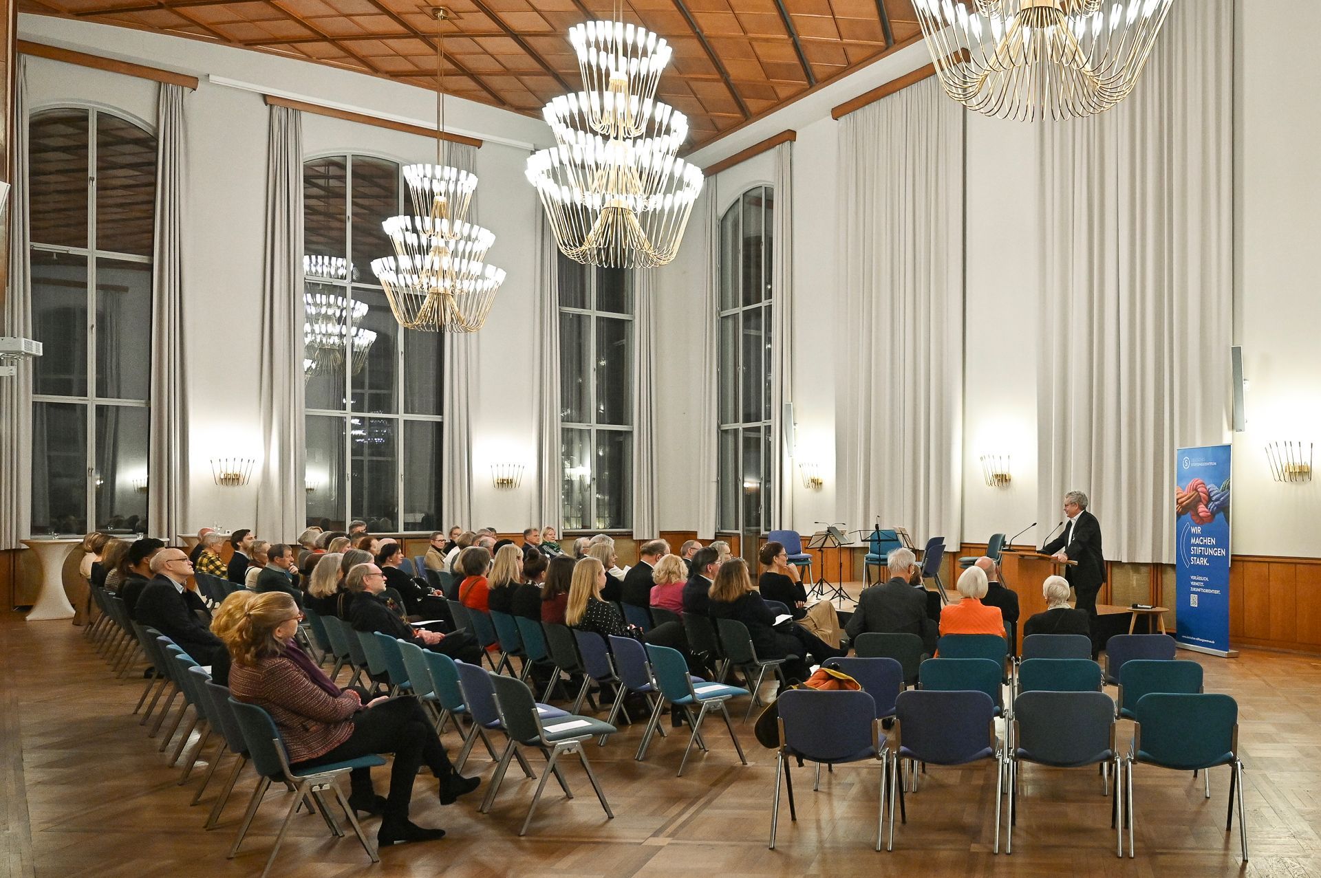 Zuschauer sitzen in einem großen Saal und hören einem Redner am Rednerpult zu. Darüber hängen Kronleuchter.