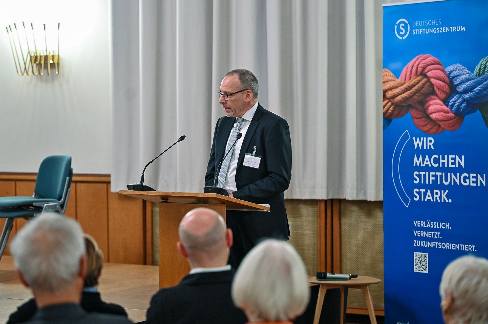 Ein Mann spricht auf einem Podium im Rahmen einer Konferenz, vor Publikum und einem Banner.